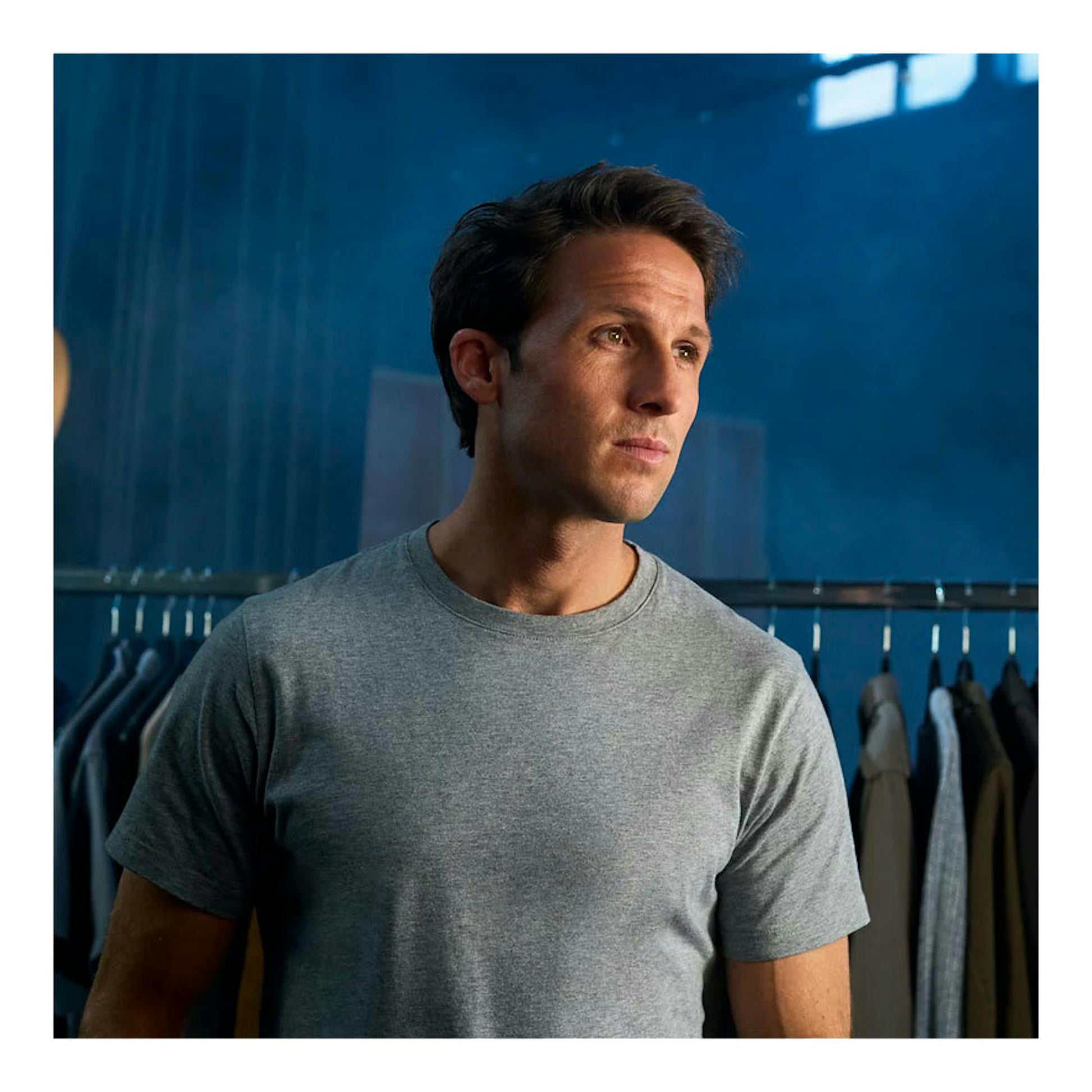 Hombre con camiseta gris, vista de tres cuartos, frente a un perchero con ropa y pared azul.
