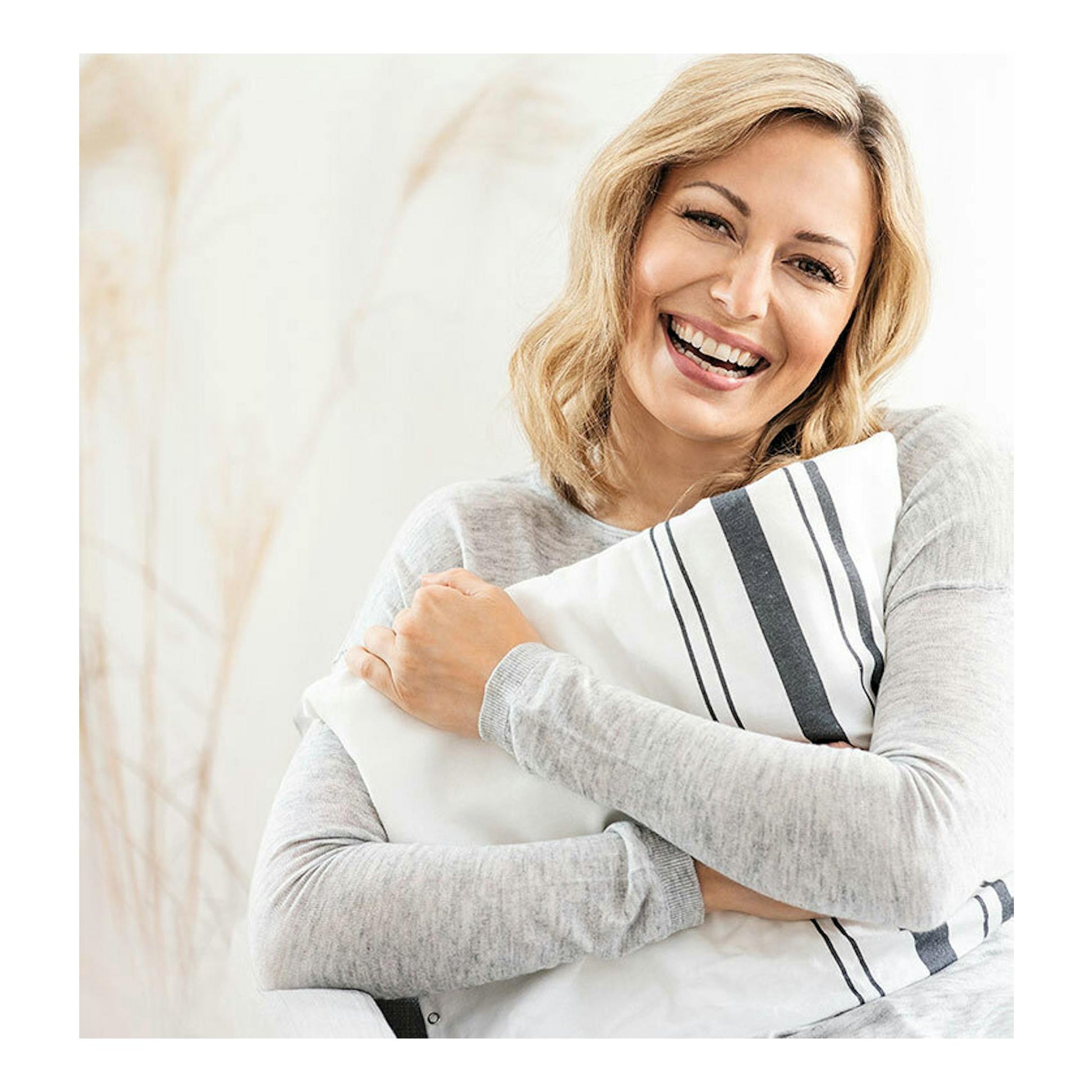 Mujer sonriente abrazando una almohadilla blanca y gris a rayas, vista frontal. Beurer HK 25