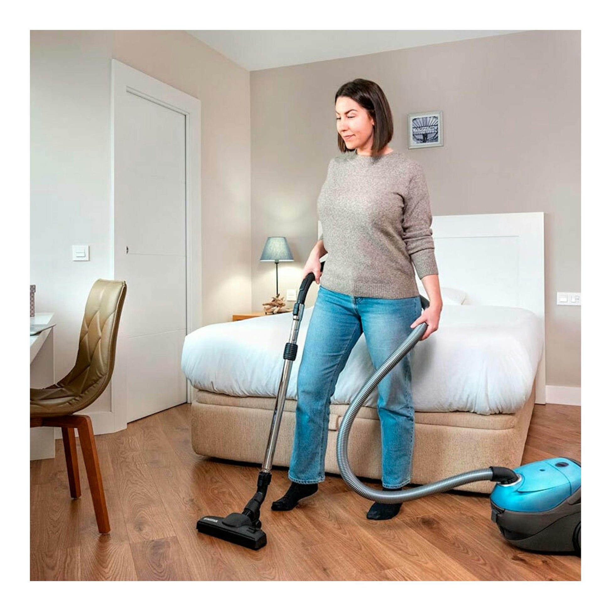 Mujer aspirando un dormitorio con aspirador Nilfisk ONE azul y gris, vista frontal.