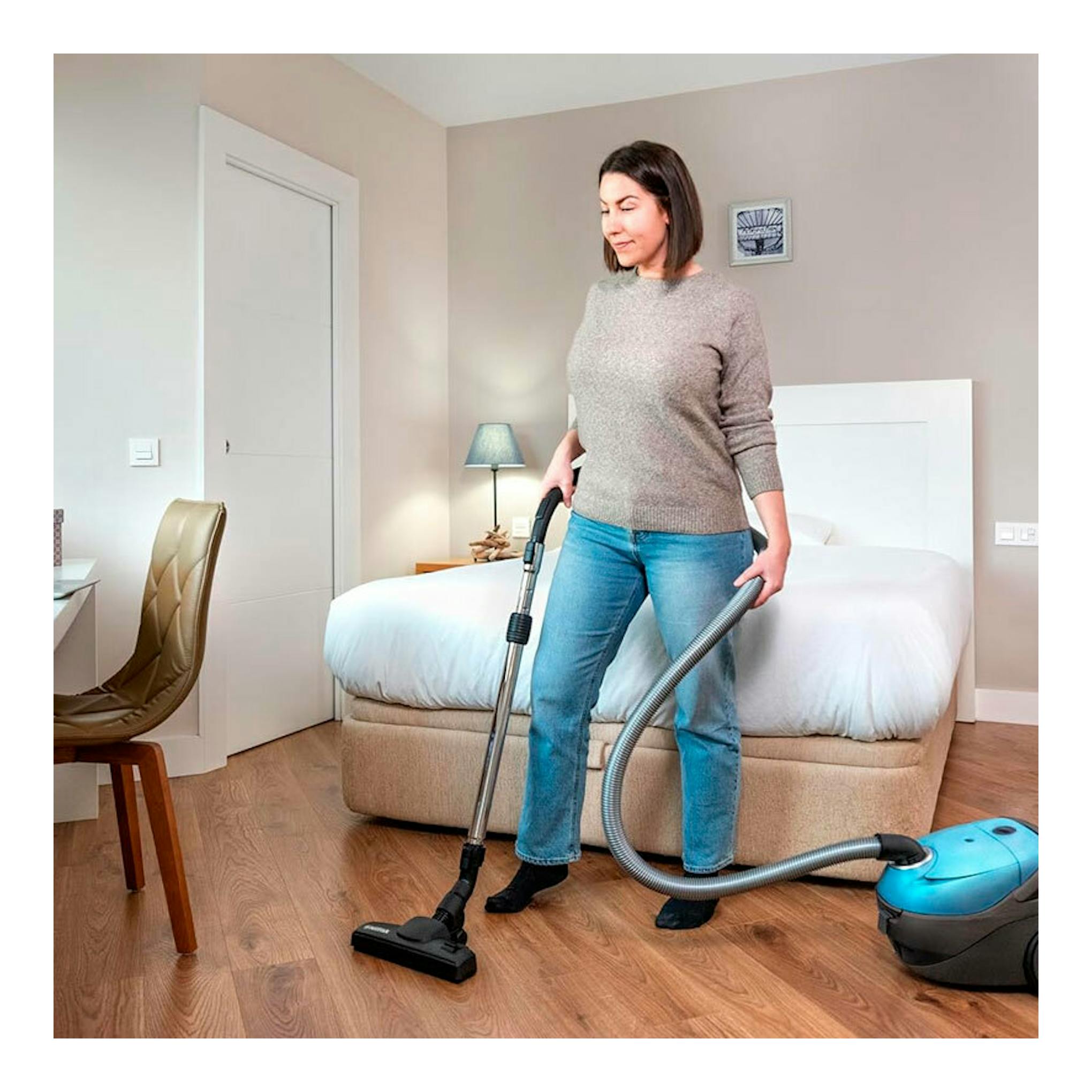 Mujer aspirando un dormitorio con aspirador Nilfisk ONE azul y gris, vista frontal.