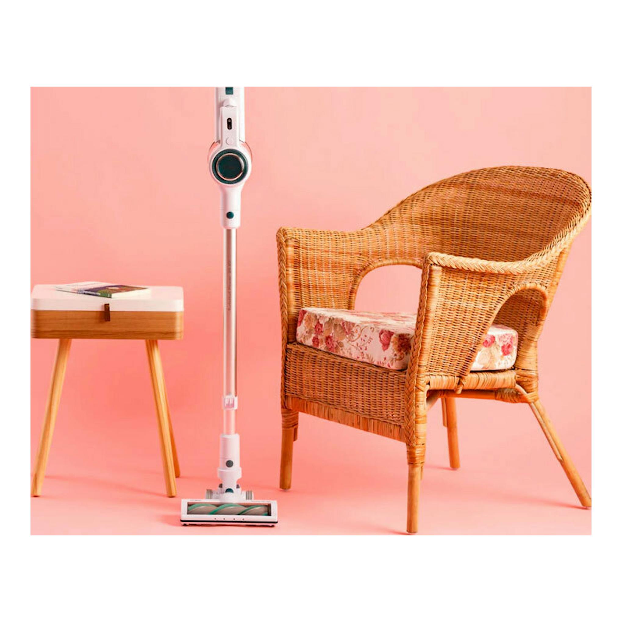 Aspirador vertical Solac AE2505 blanco, de pie sobre fondo rosa, junto a una silla de mimbre y una mesa auxiliar.