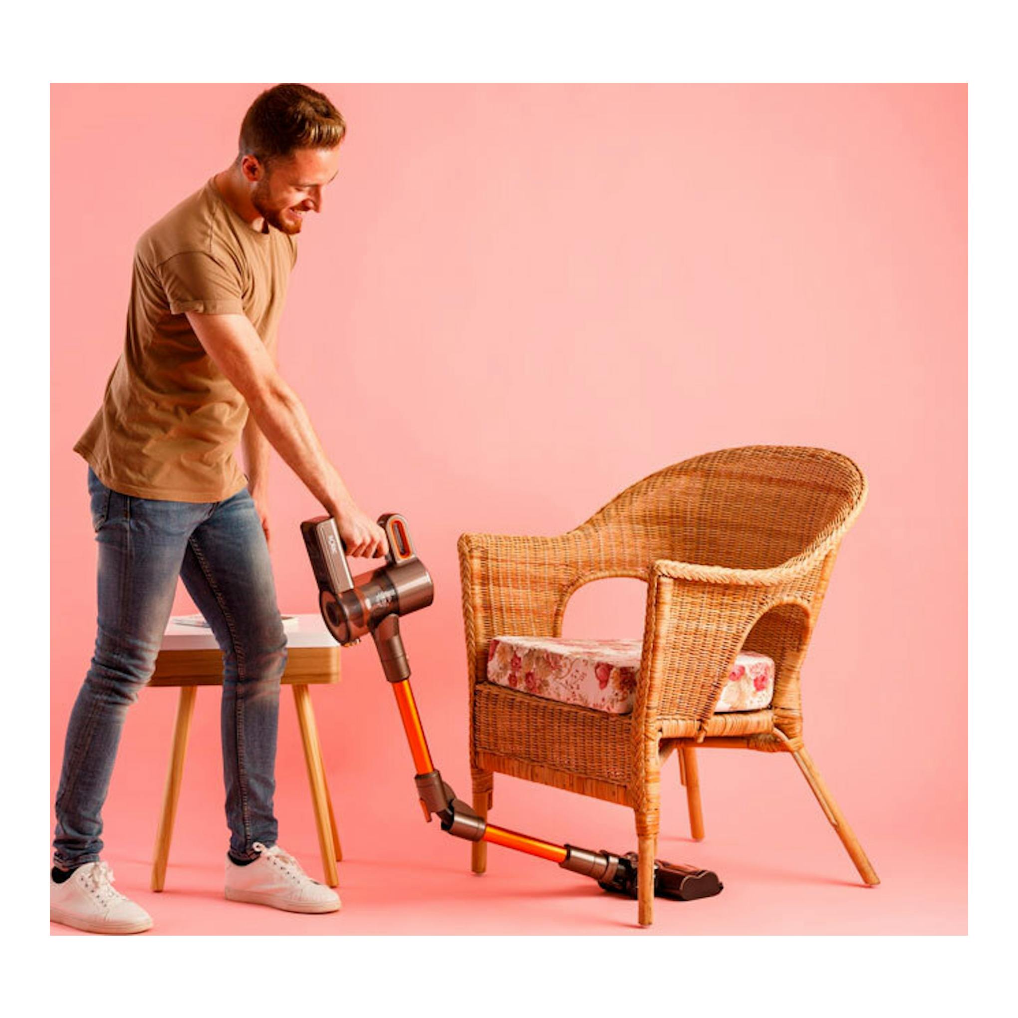 Solac AE2515: aspirador sin cable naranja y negro; un hombre limpia una silla de mimbre sobre fondo rosa.