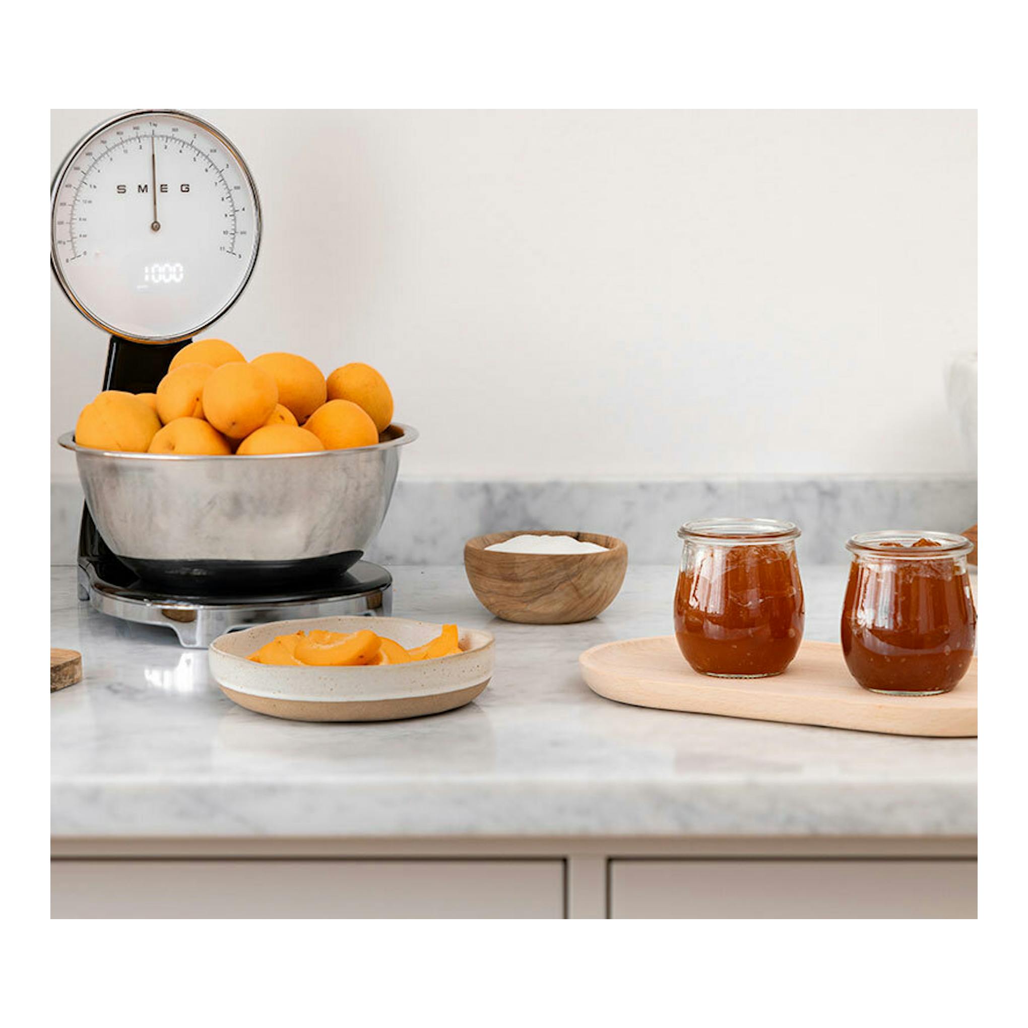 Balanza de cocina Smeg KSF01BLWW negra, vista frontal, con bol metálico lleno de naranjas sobre encimera.