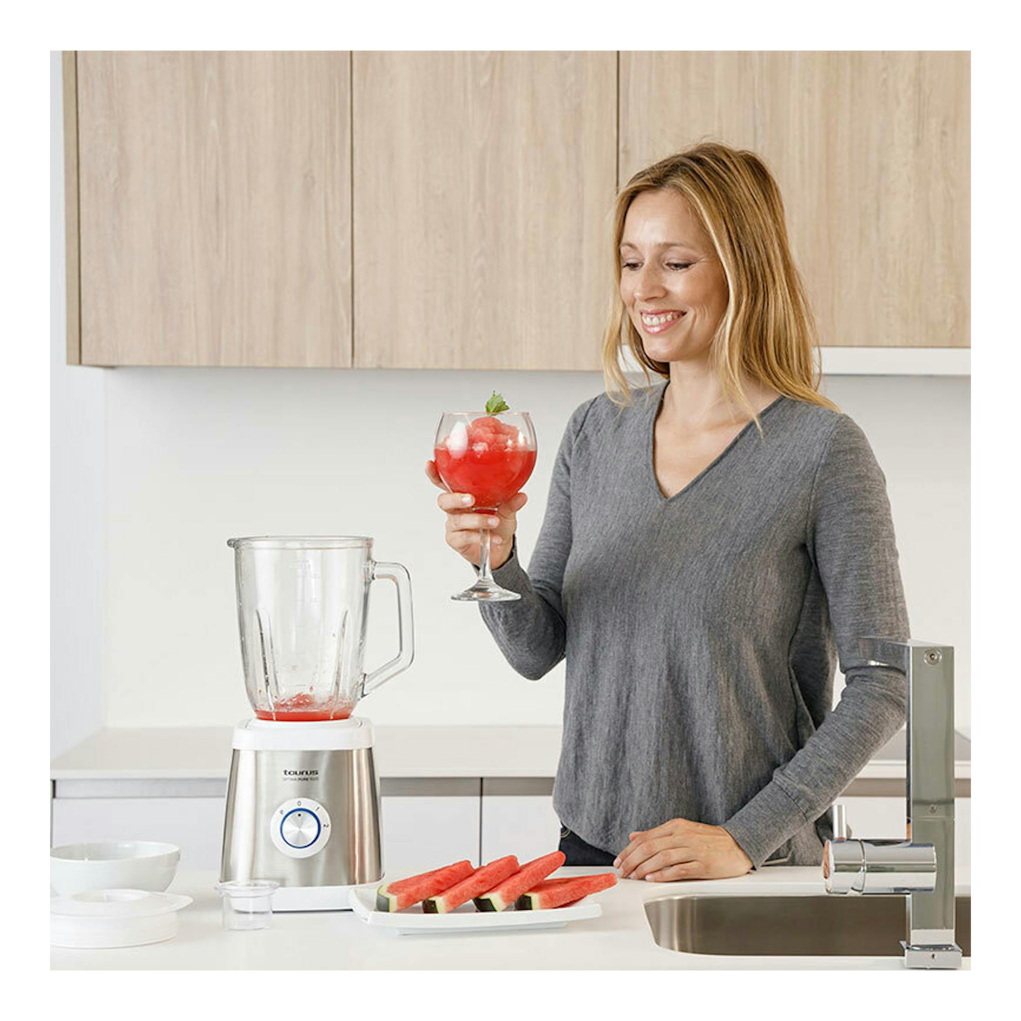 Batidora de vaso Taurus Optima Pure 1500 plateada sobre encimera, junto a una mujer con una copa roja en cocina.