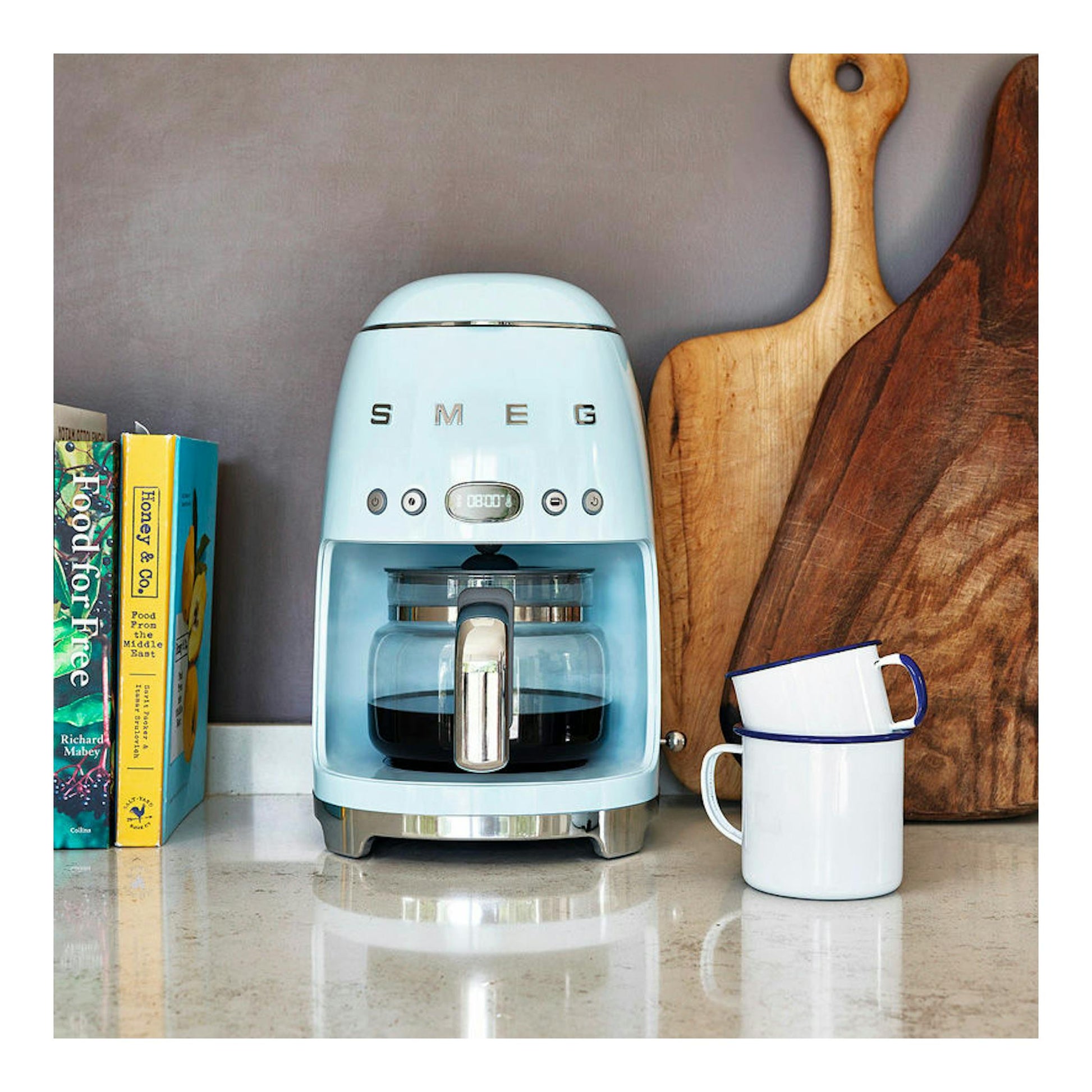 Cafetera de goteo Smeg DCF02PBEU azul pastel, vista frontal, sobre encimera con taza y tablas de madera.
