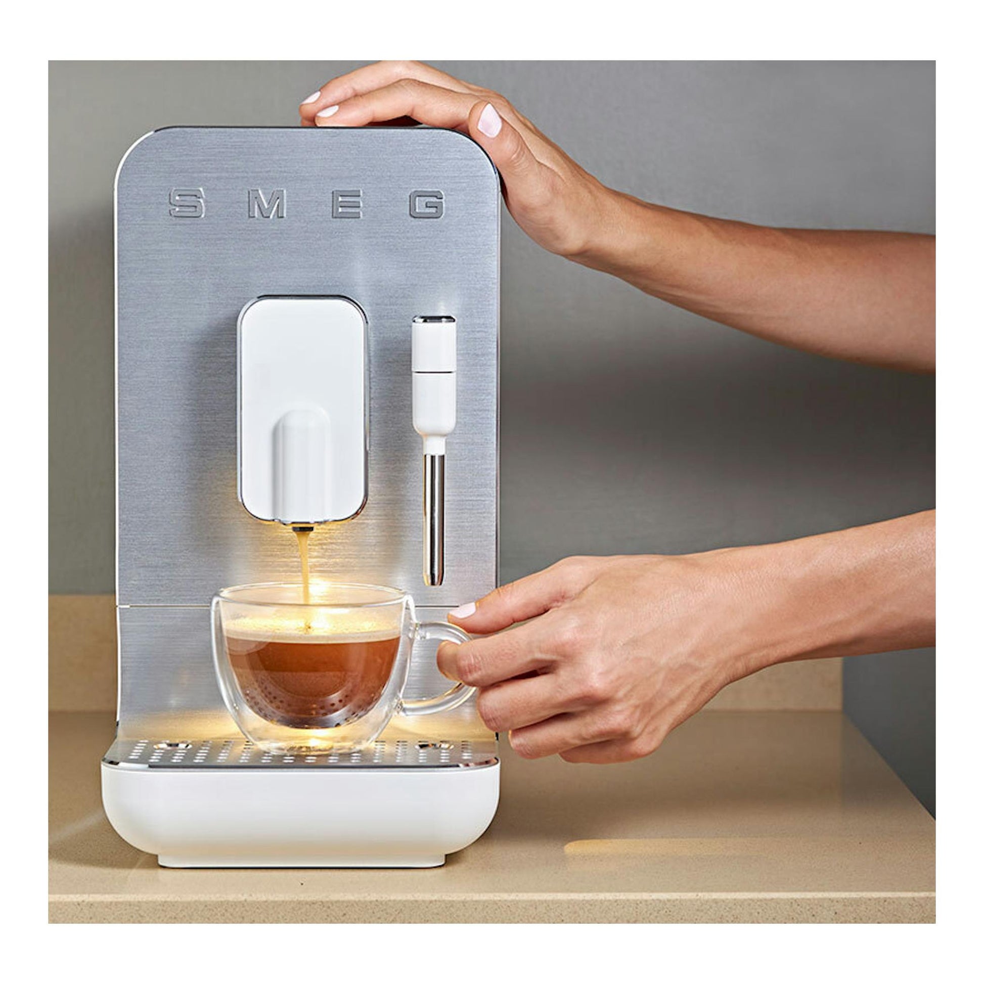Cafetera superautomática Smeg, vista frontal, en acero inoxidable, sirviendo café en una taza de vidrio.