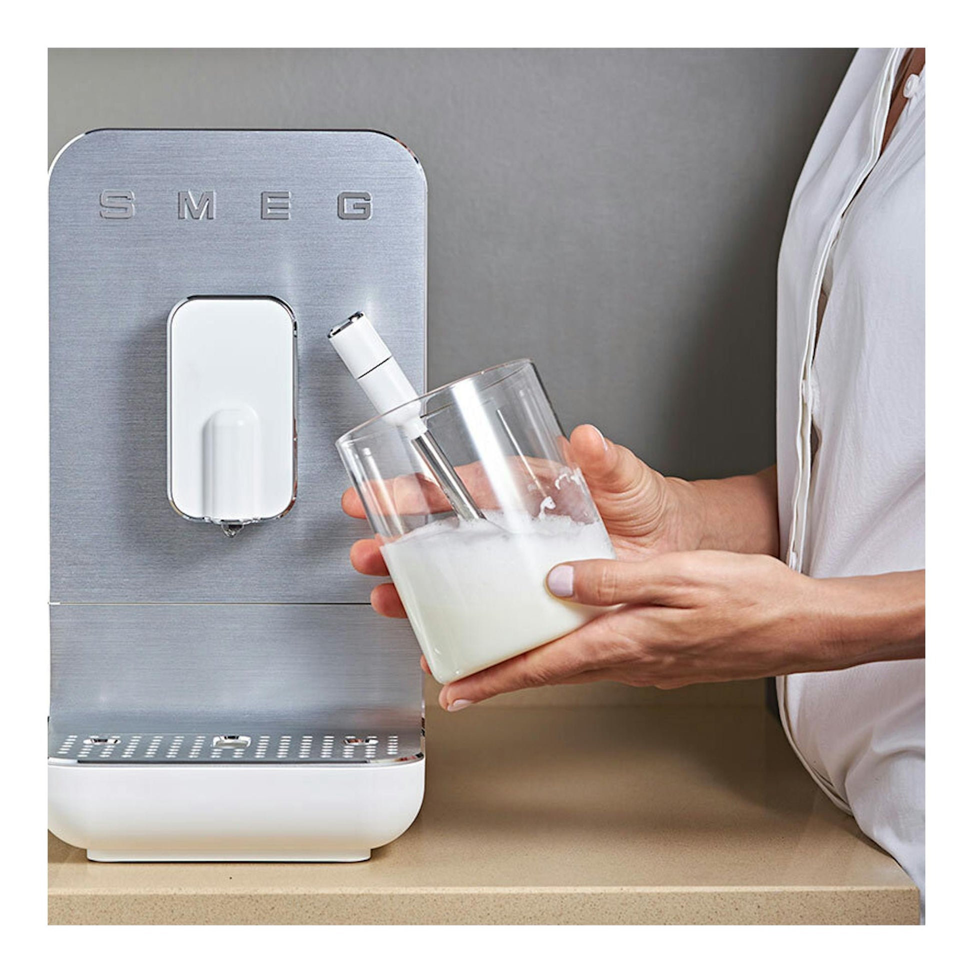 Cafetera superautomática Smeg BCC12WHMEU, vista frontal, en blanco y acero, con una mano sosteniendo un vaso.