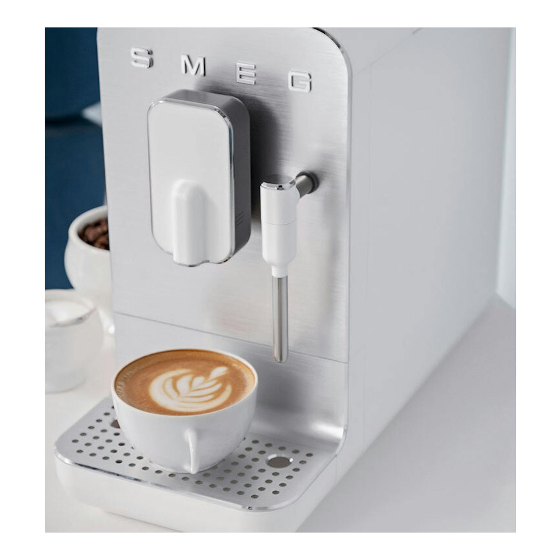Cafetera superautomática Smeg BCC12WHMEU blanca y plateada, vista frontal en ángulo, con taza de café.