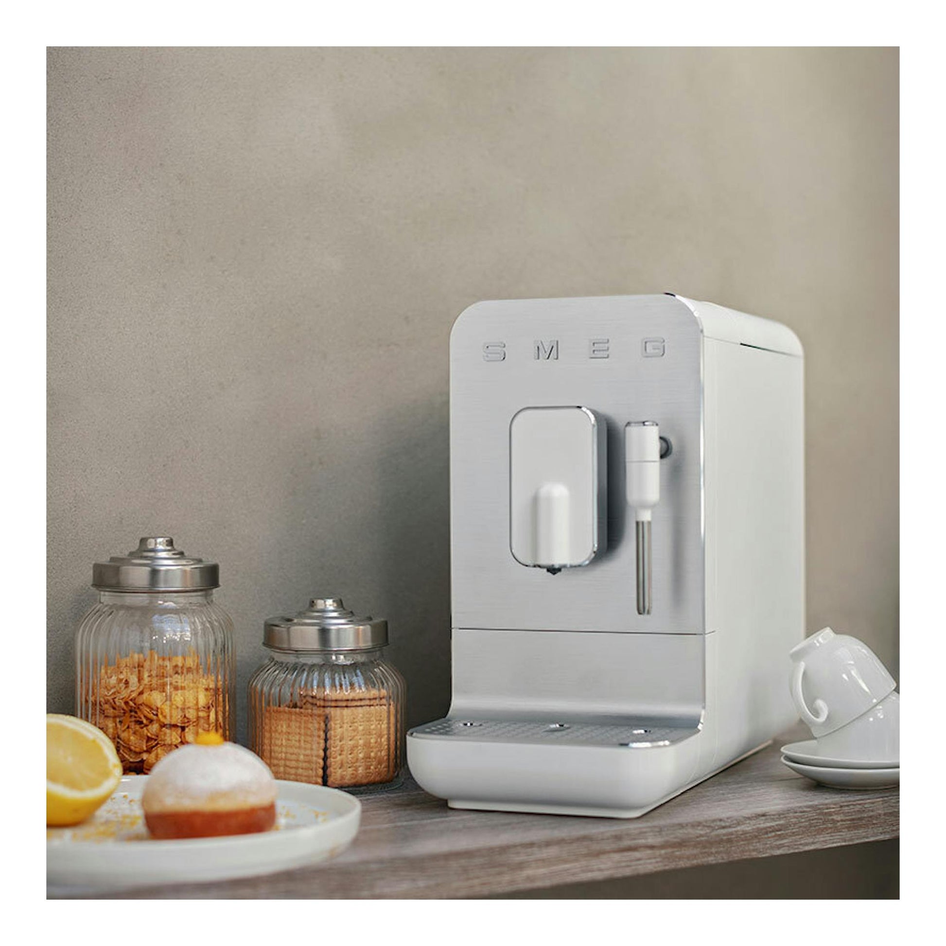 Cafetera superautomática Smeg BCC12WHMEU blanca, vista frontal en ángulo, sobre una mesa junto a tazas y tarros.
