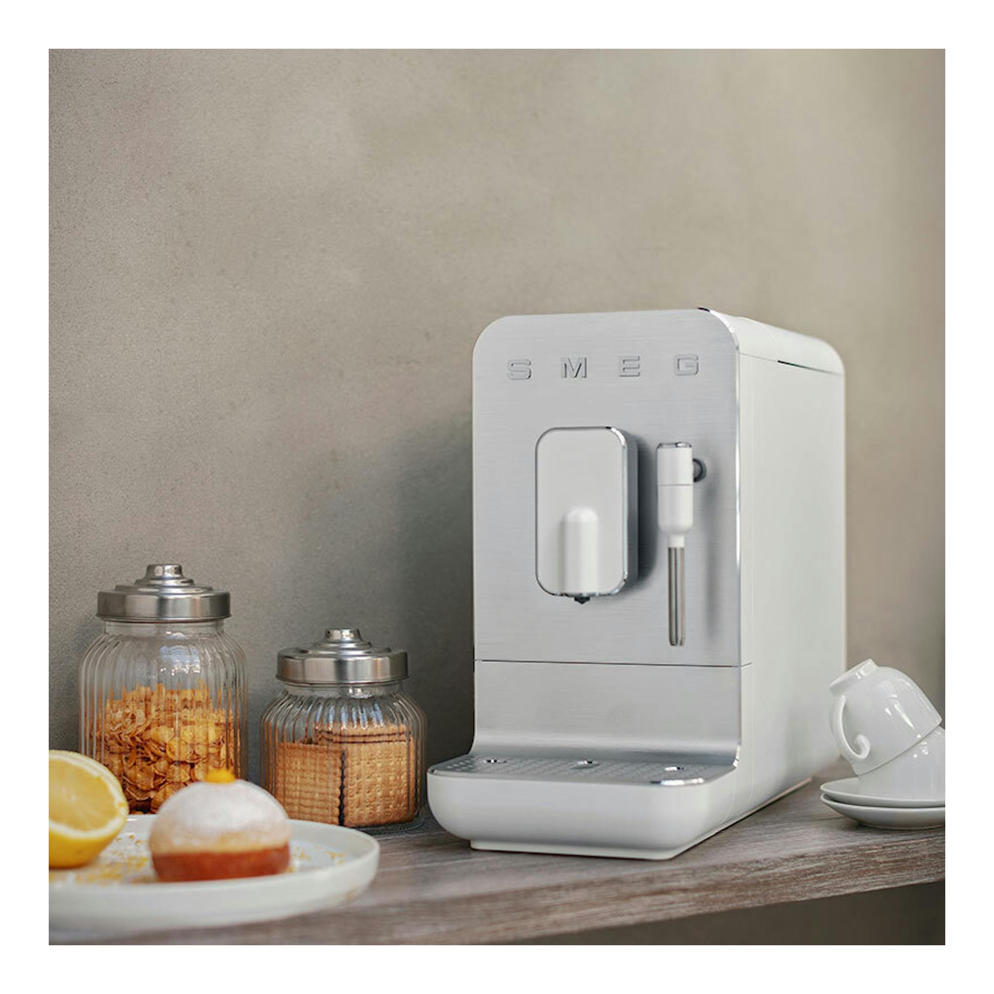 Cafetera superautomática Smeg BCC12WHMEU blanca, vista frontal en ángulo, sobre una mesa junto a tazas y tarros.