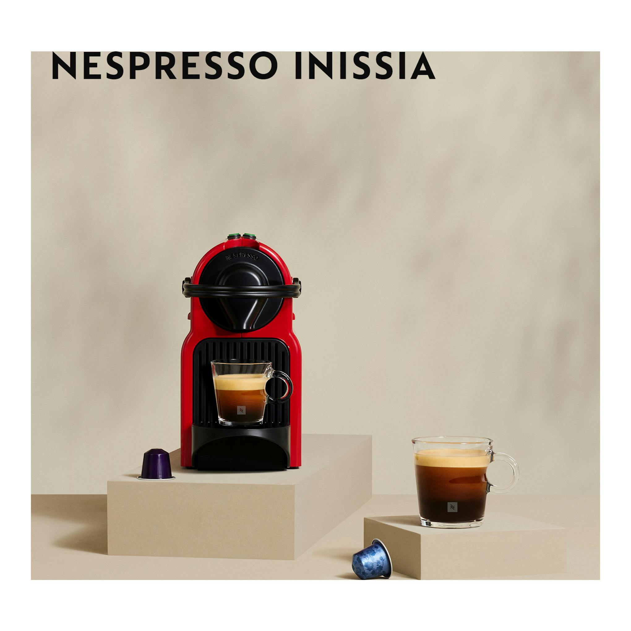 Cafetera de cápsulas Nespresso Inissia roja, vista frontal, con taza de café y cápsulas sobre pedestal.