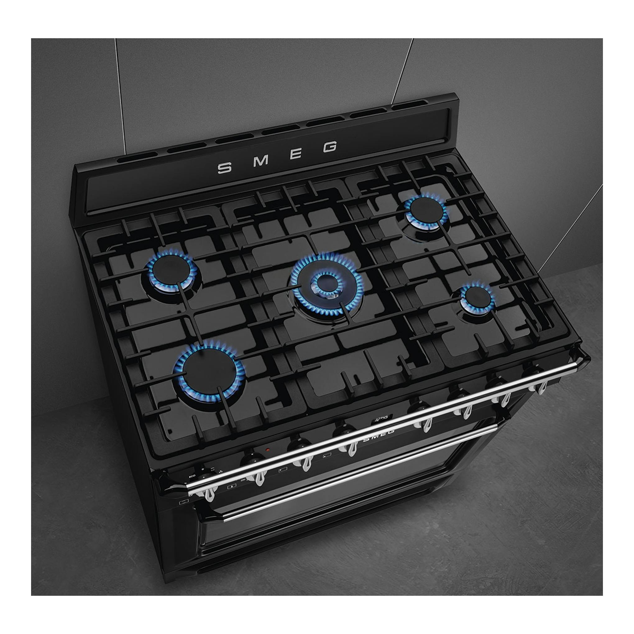 Cocina de gas Smeg TR90GMBL negra, vista en ángulo frontal, con 5 quemadores encendidos.