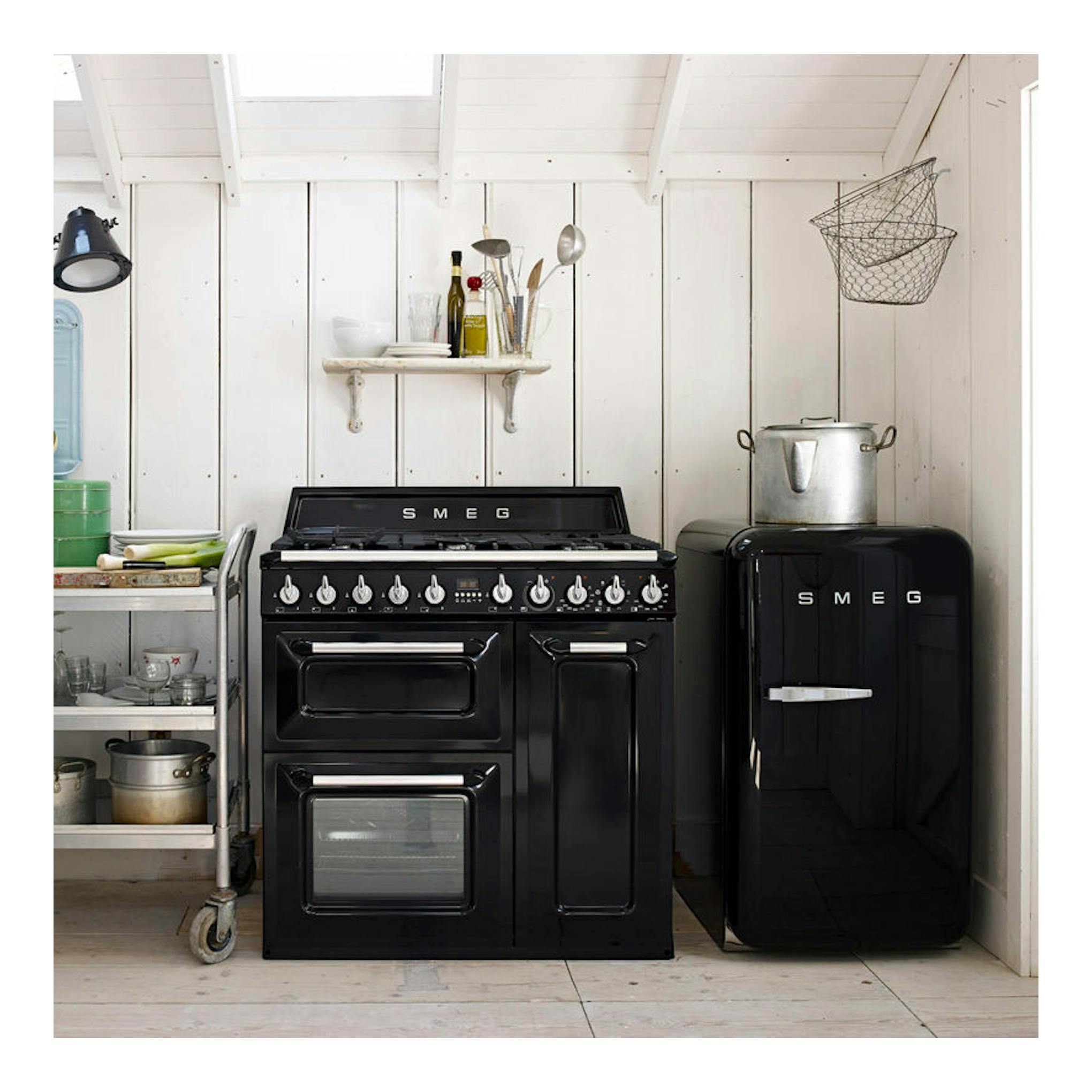 Cocina Smeg TR93BL negra de 90 cm, vista frontal, con horno doble y junto a un frigorífico Smeg negro.