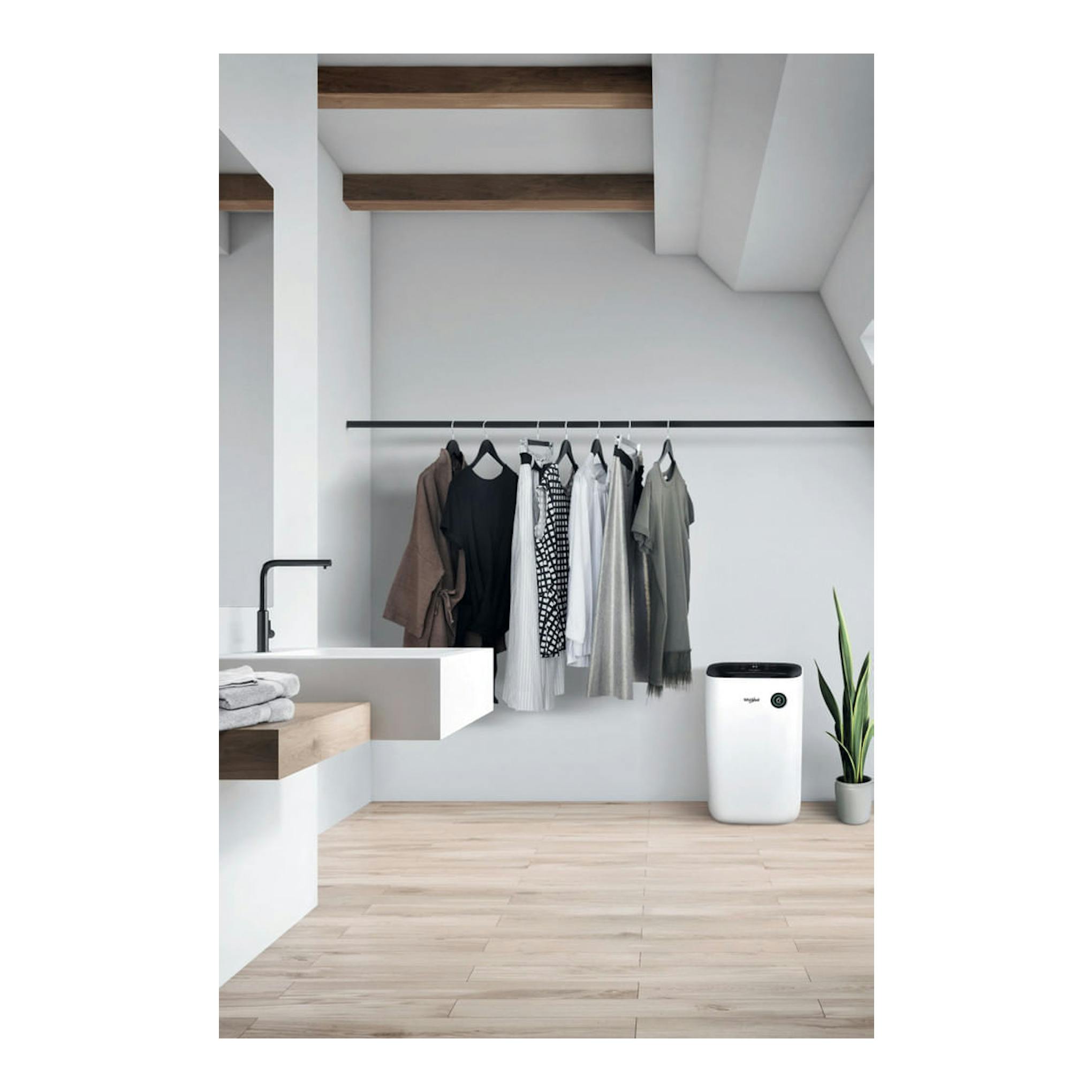 Deshumidificador Whirlpool DE20W5252 blanco, vista frontal, junto a ropa colgada y una planta en una estancia.