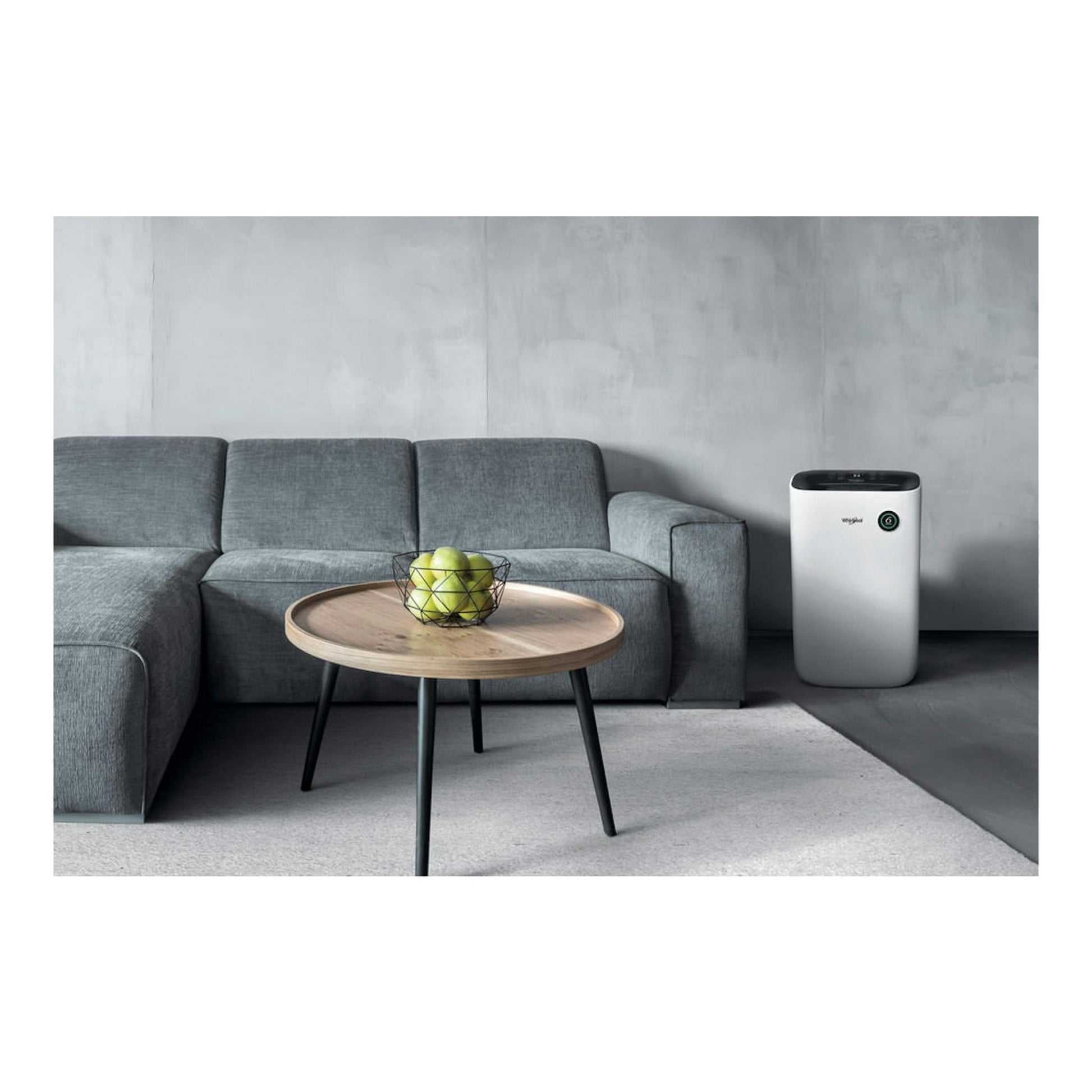 Deshumidificador Whirlpool blanco, vista frontal, junto a un sofá gris y una mesa redonda en un salón.