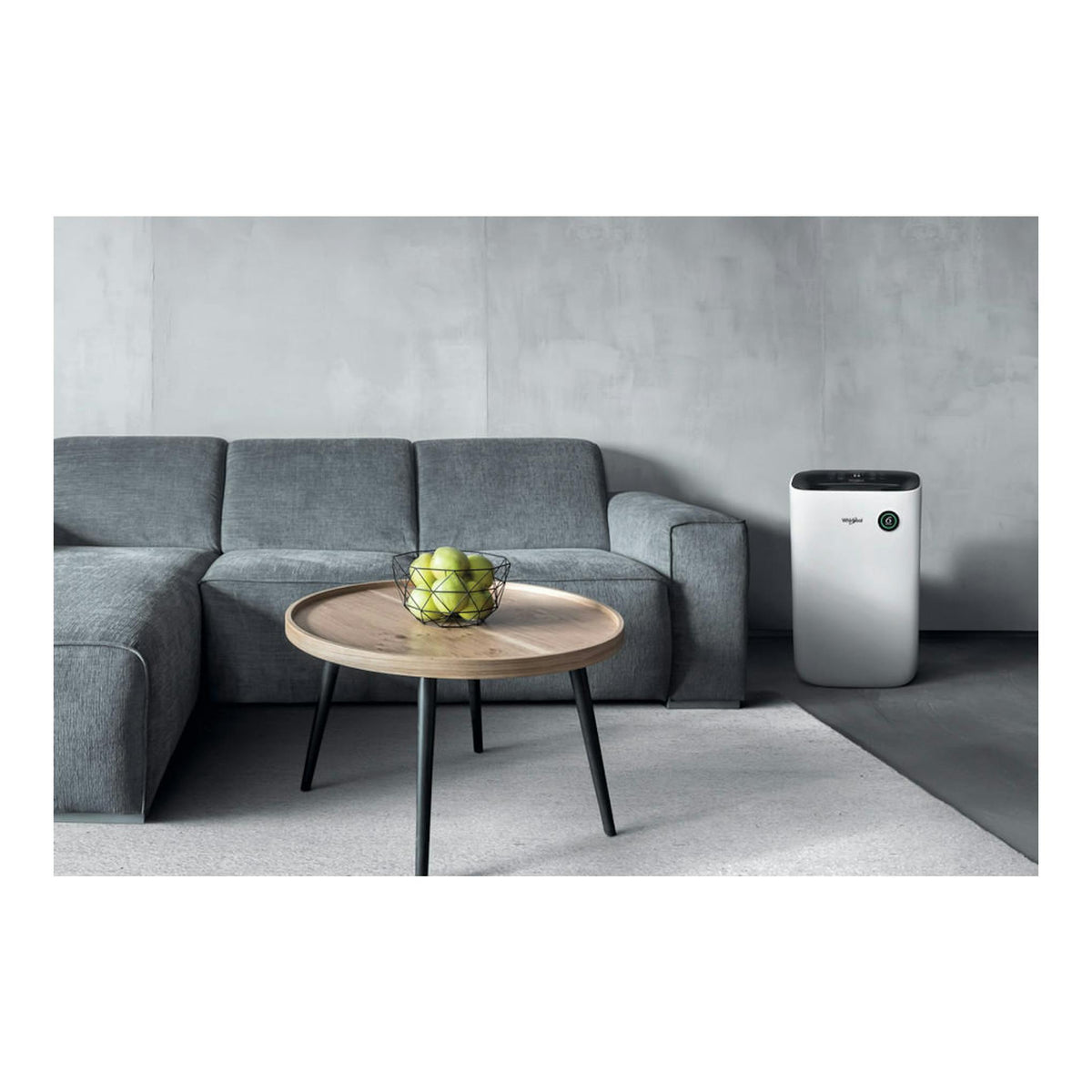 Deshumidificador Whirlpool blanco, vista frontal, junto a un sofá gris y una mesa redonda en un salón.