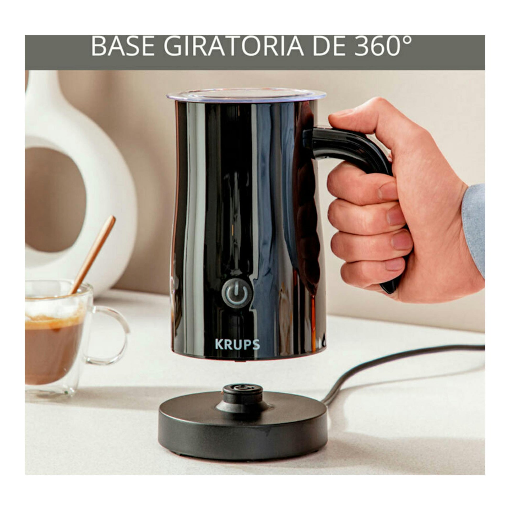 Espumador de leche Krups negro, sostenido con la mano sobre base giratoria de 360°, con taza al lado.
