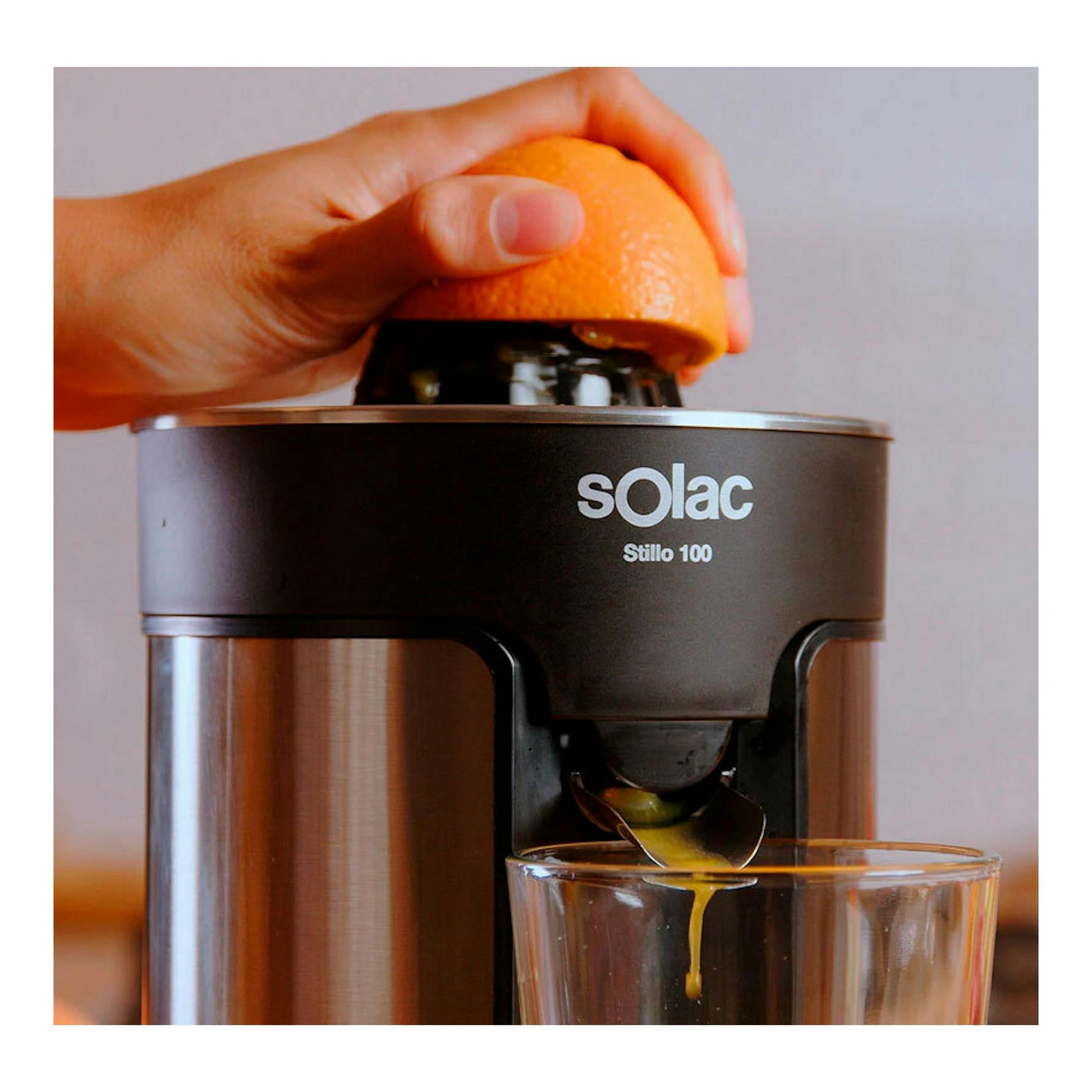 Exprimidor Solac Stillo 100 negro, vista frontal, con una mano exprimiendo una naranja y zumo saliendo al vaso.