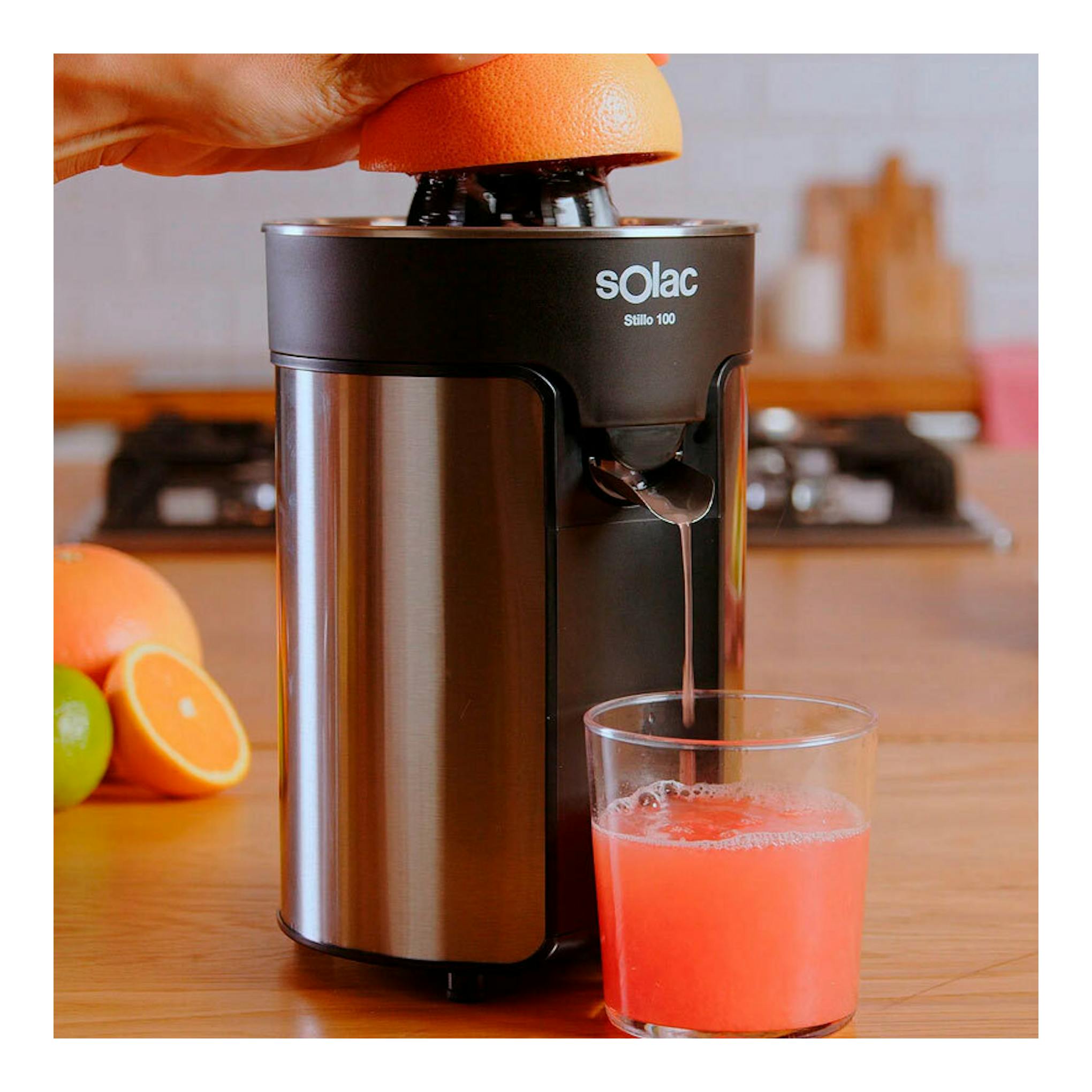 Exprimidor eléctrico Solac Stillo 100 negro y acero, con una naranja prensada y un vaso de zumo sobre una encimera.