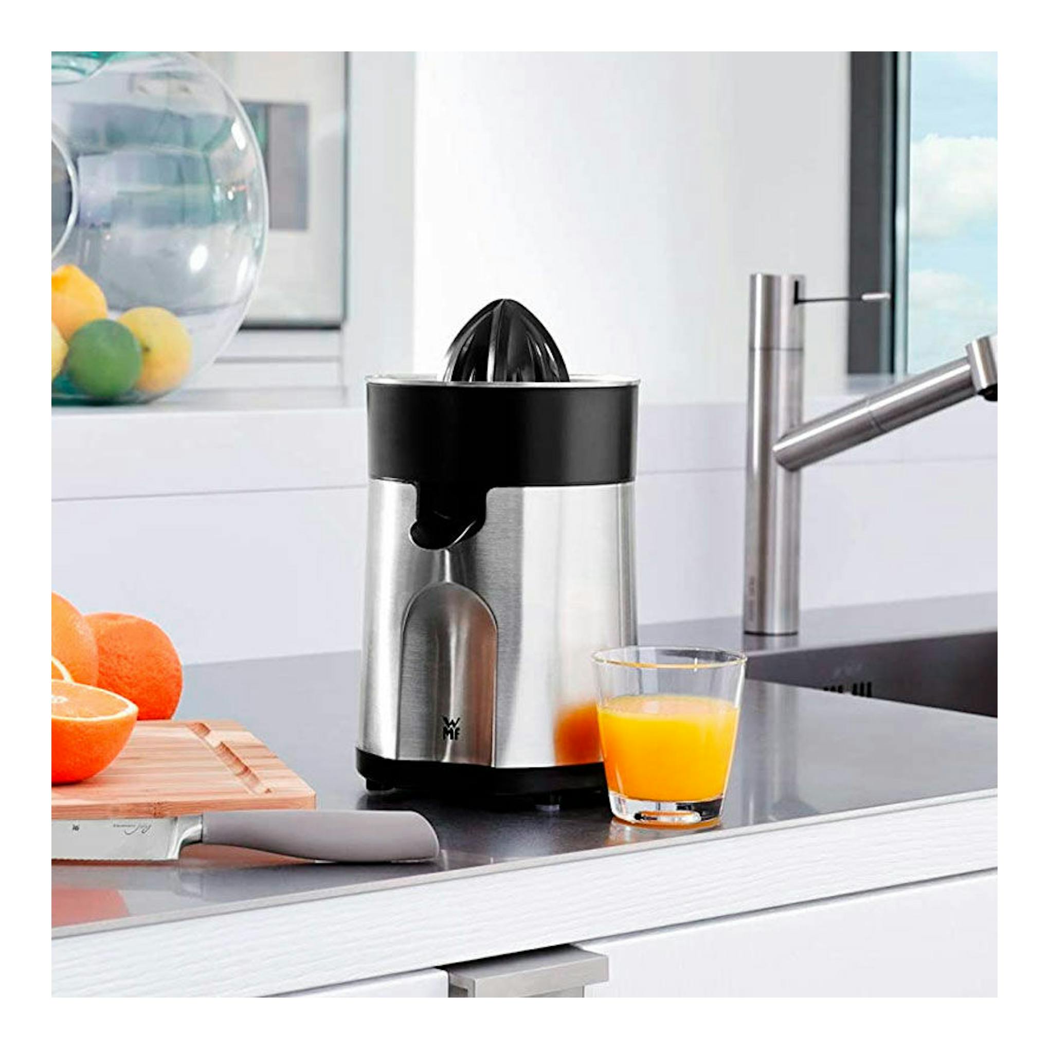Exprimidor WMF STELIO de acero inoxidable y negro, vista frontal en encimera con vaso de zumo al lado.