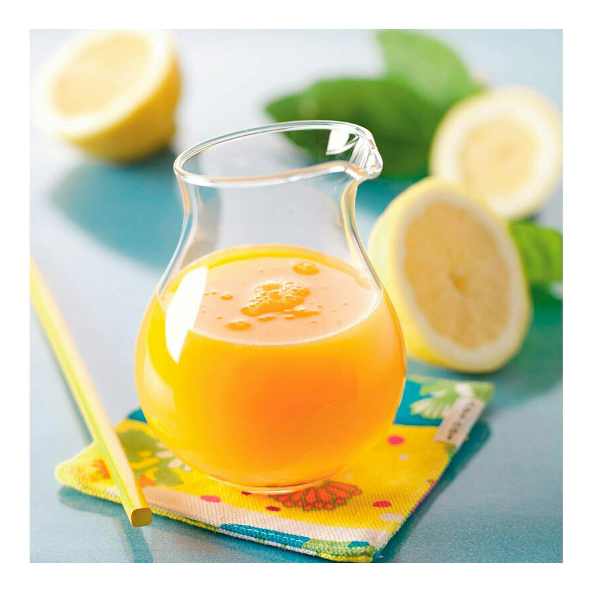 Jarra de vidrio con zumo de naranja sobre un posavasos, con limones cortados y hojas verdes al fondo.