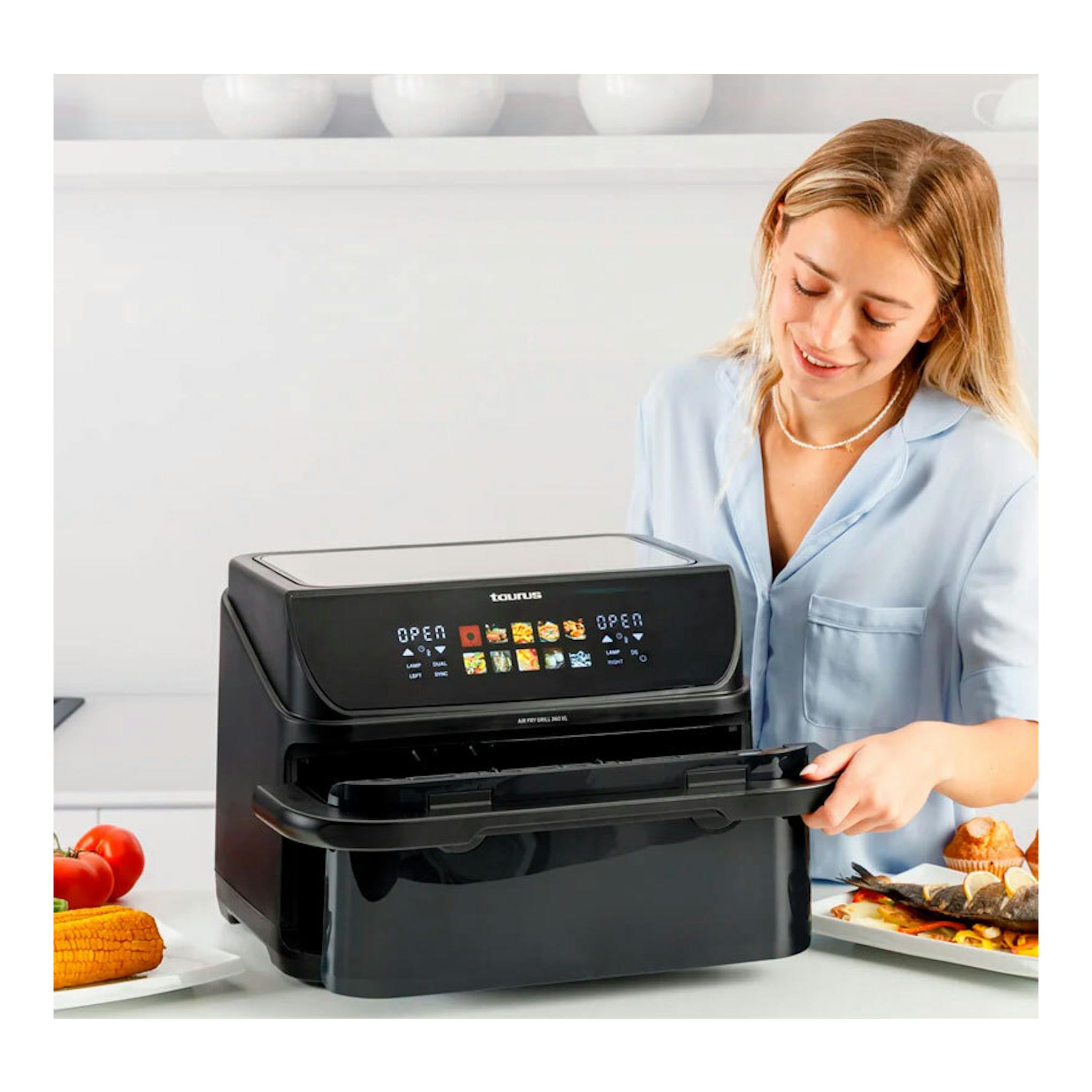Freidora de aire Taurus Grill 360 negra, vista frontal, con una mujer abriendo la bandeja en una cocina.