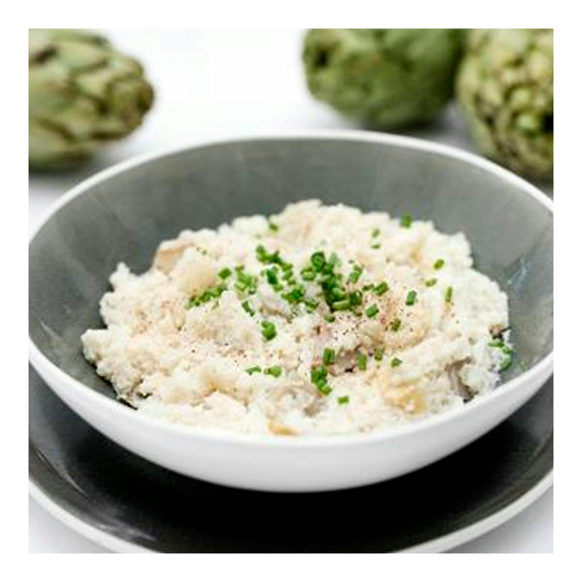 Bol de cerámica gris con arroz cremoso y cebollino, sobre plato negro, con alcachofas desenfocadas al fondo.