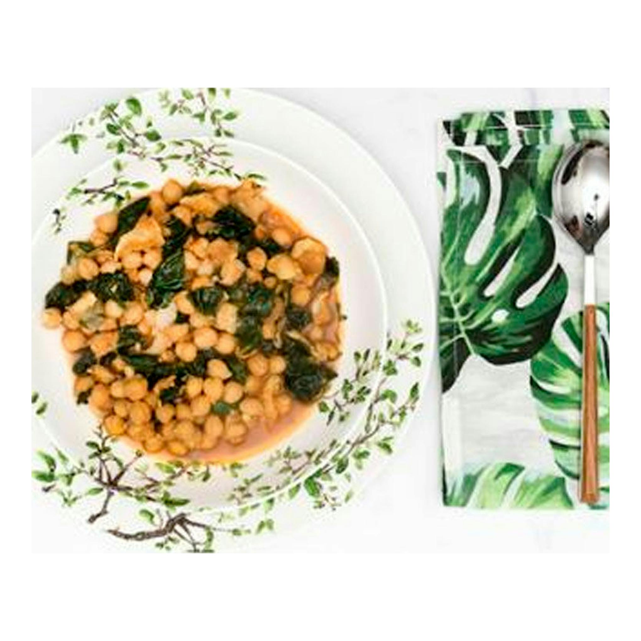 Plato blanco con garbanzos y verduras sobre mesa blanca, junto a servilleta verde con cubiertos.