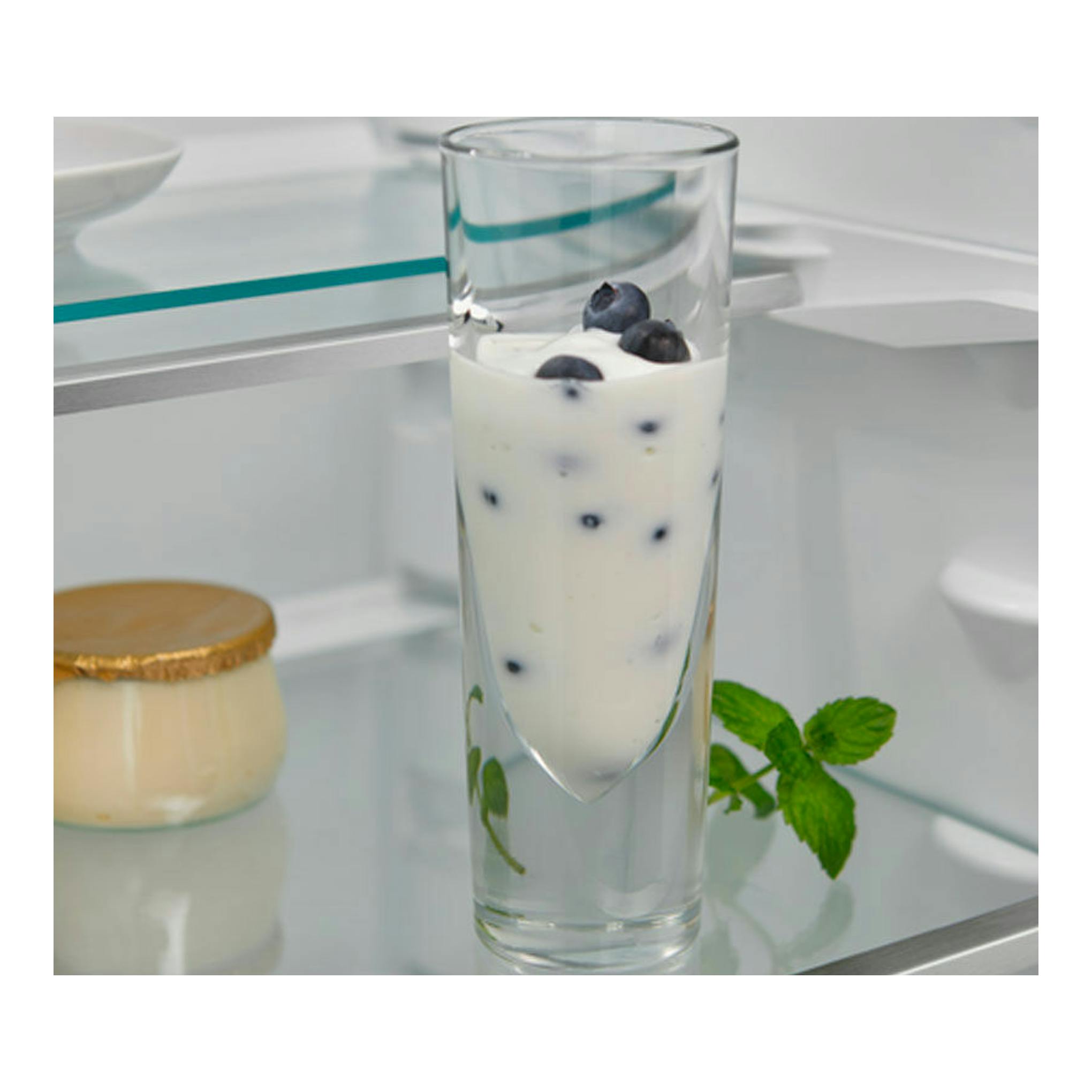 Interior de frigorífico blanco con vaso alto de postre con crema y arándanos, tarro pequeño y hojas de menta.