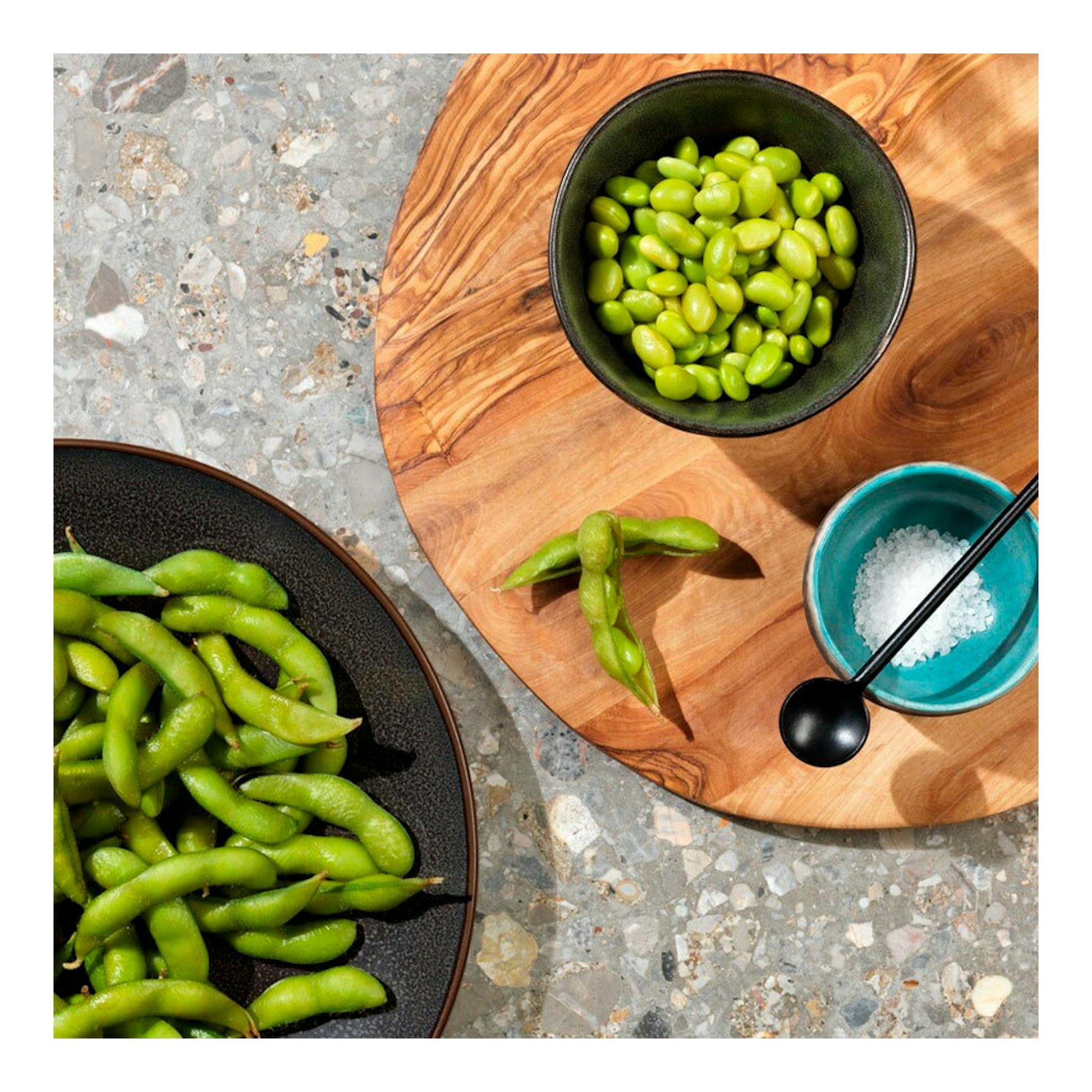Vista cenital de cuencos con edamame verde y sal sobre tabla de madera.