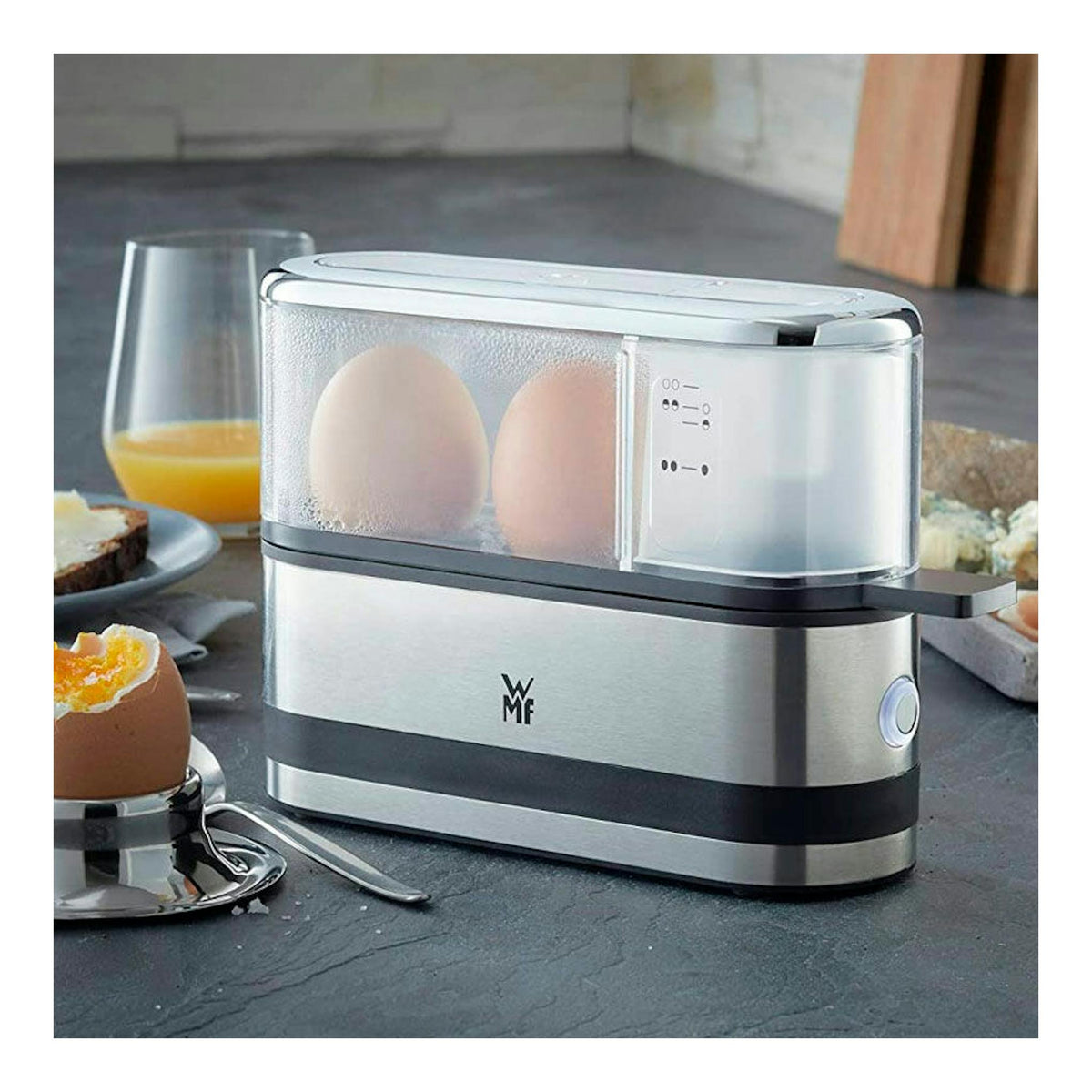 Cocedor de huevos WMF de acero inoxidable y plástico, vista frontal, con dos huevos dentro sobre una encimera.