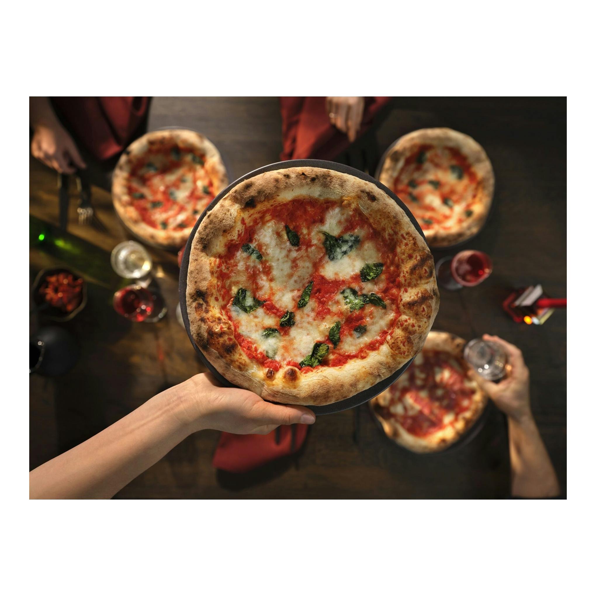 Vista cenital de varias pizzas con mozzarella y albahaca sobre una mesa, una en primer plano sobre una mano.