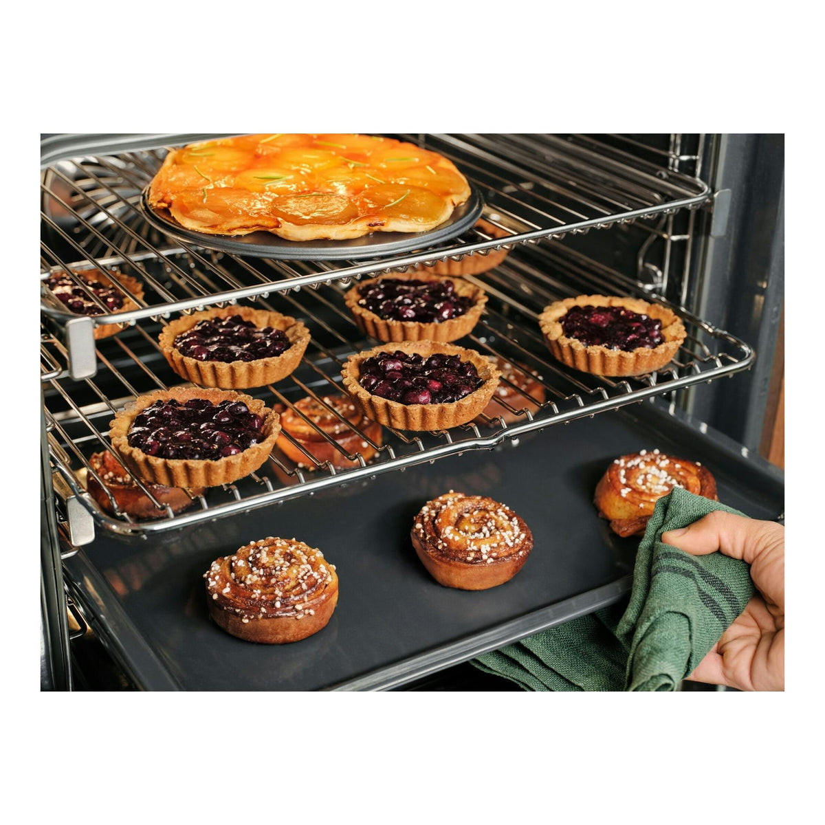 Interior de horno Electrolux COP846X1 con tartaletas y bollos; mano con paño verde.