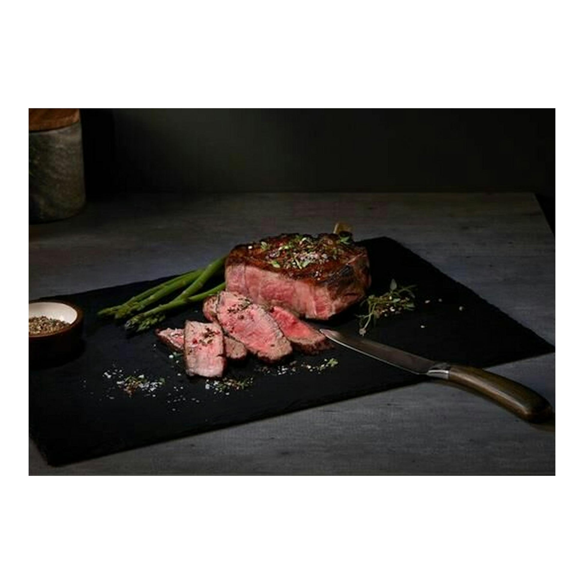 Filete de ternera cortado sobre tabla negra con espárragos, cuchillo y condimentos.