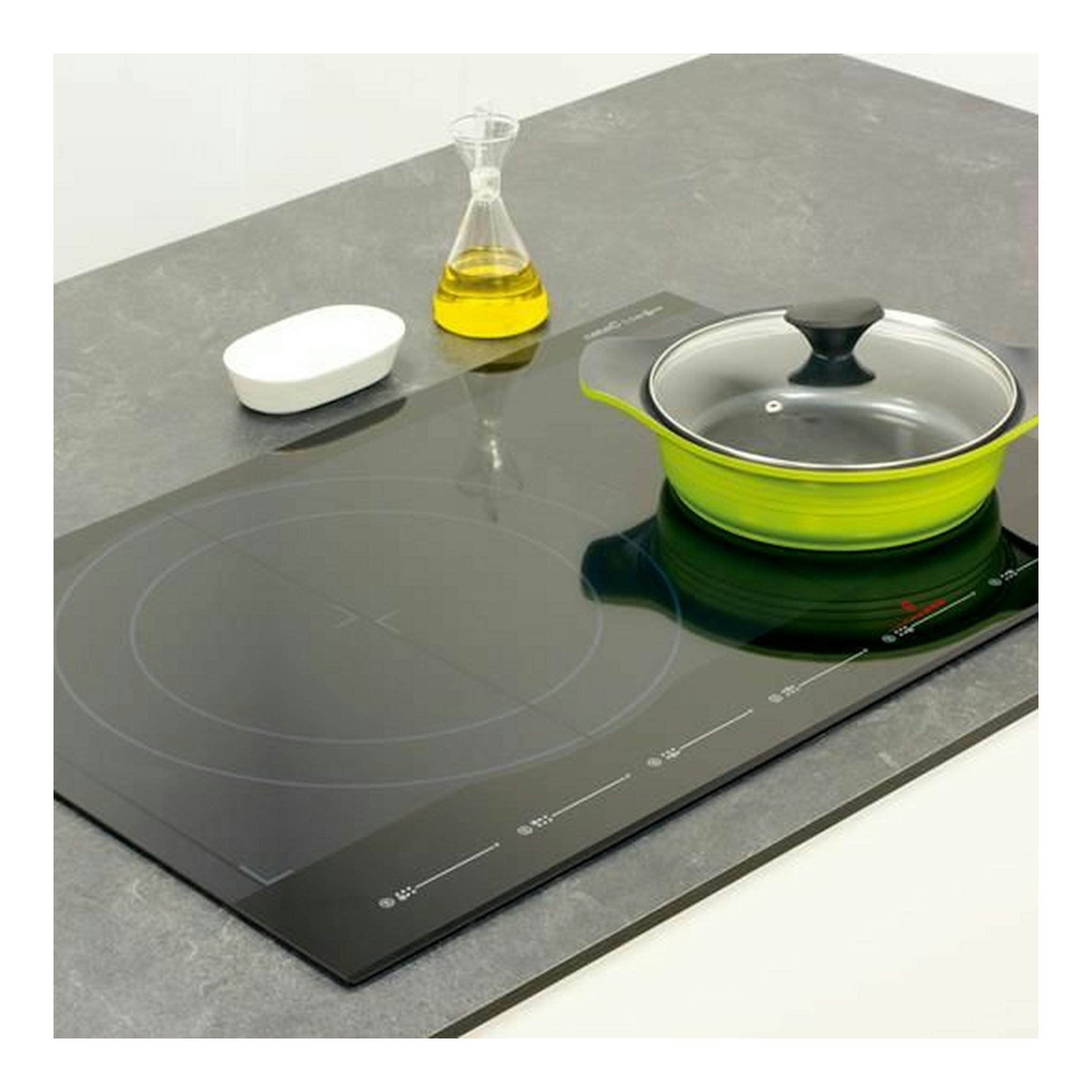 Placa de inducción Cata Giga750BK negra, vista en ángulo, con cazo verde y tapa de vidrio sobre encimera.