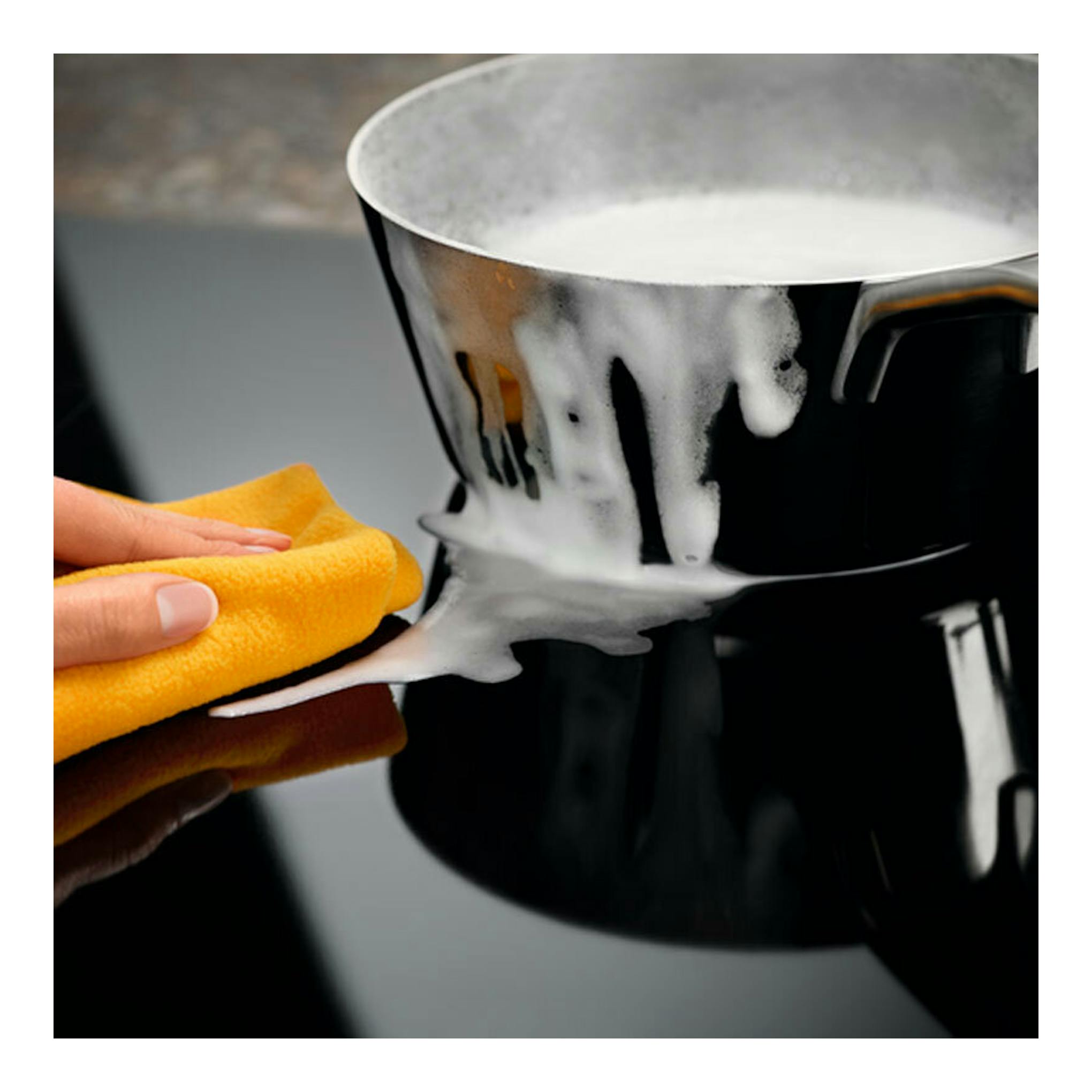 Placa de inducción Electrolux EIV95550 negra, vista en primer plano, con una olla y una mano limpiando espuma con un paño.