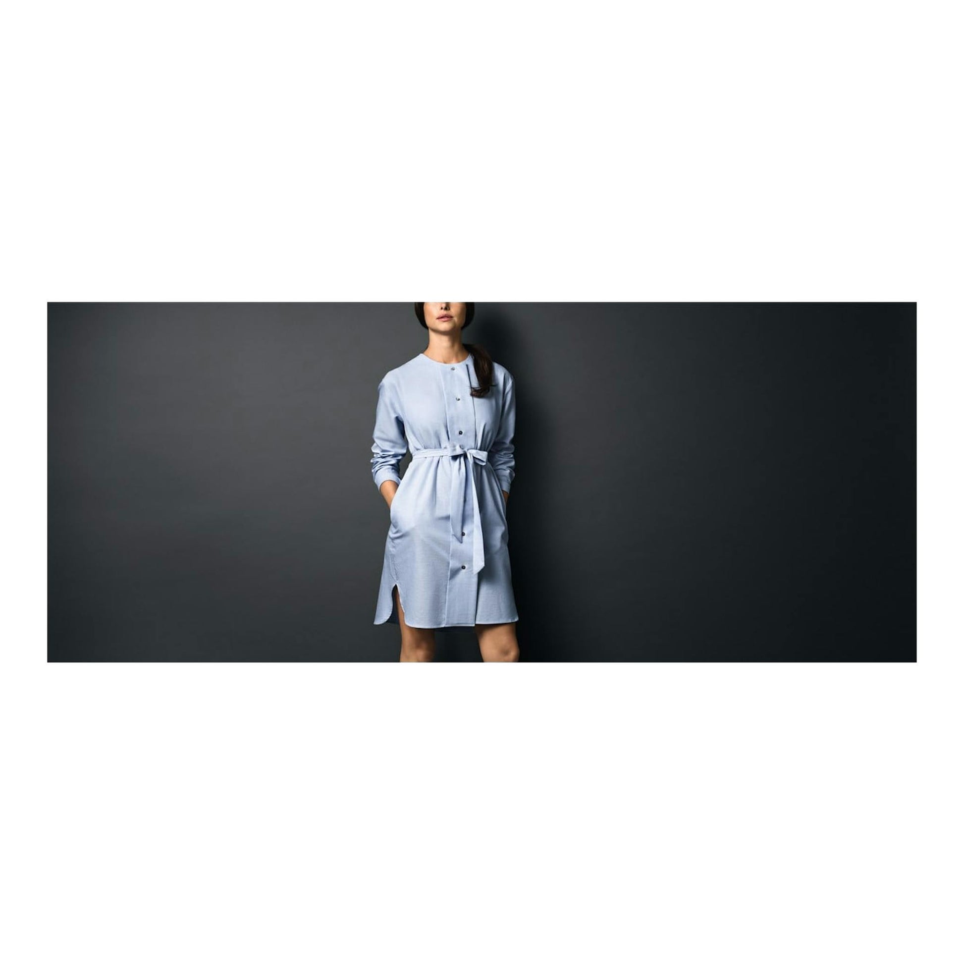 Mujer de pie con vestido camisero azul claro y cinturón, sobre fondo gris oscuro, vista frontal recortada.