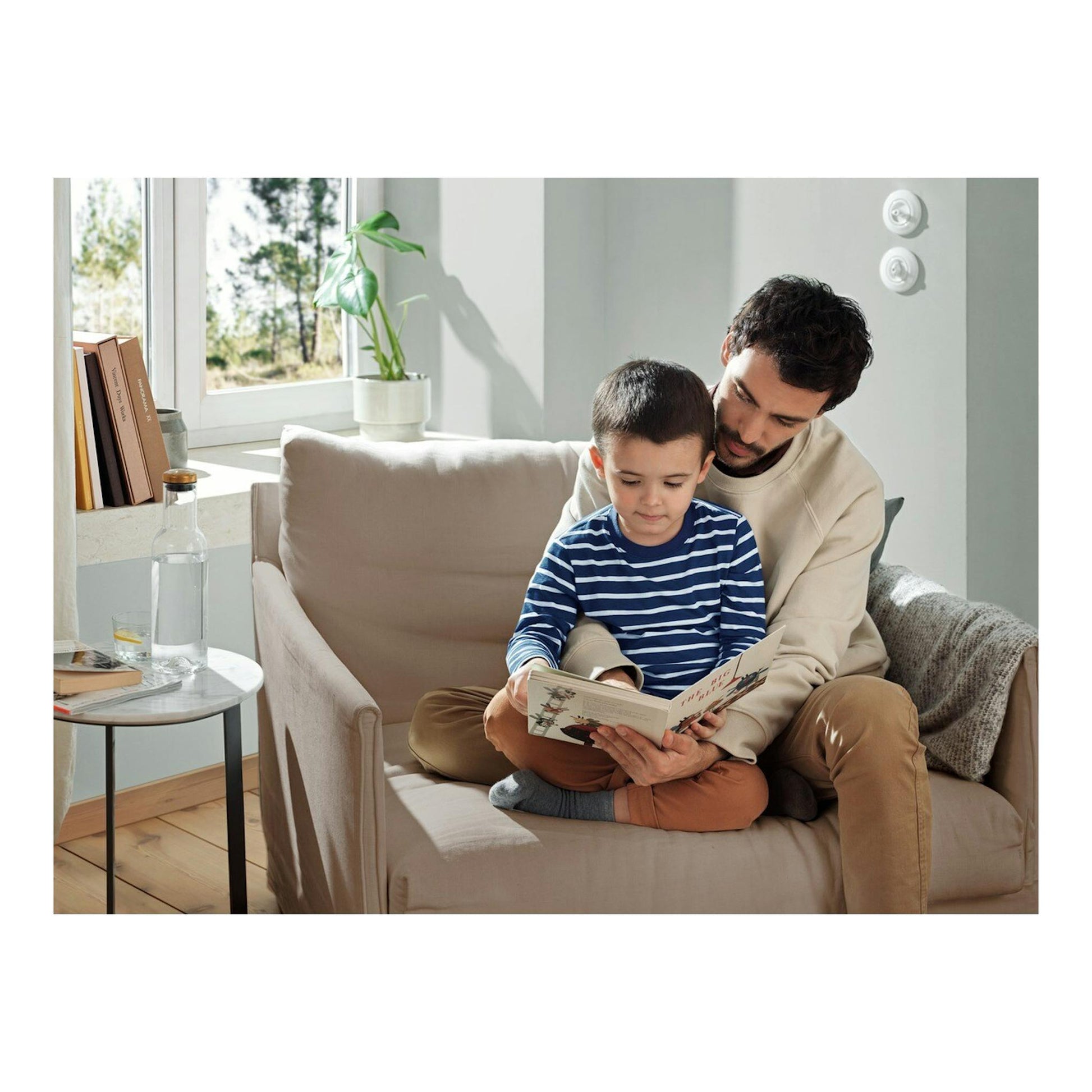 Padre e hijo sentados en un sofá leyendo un libro en un salón luminoso.