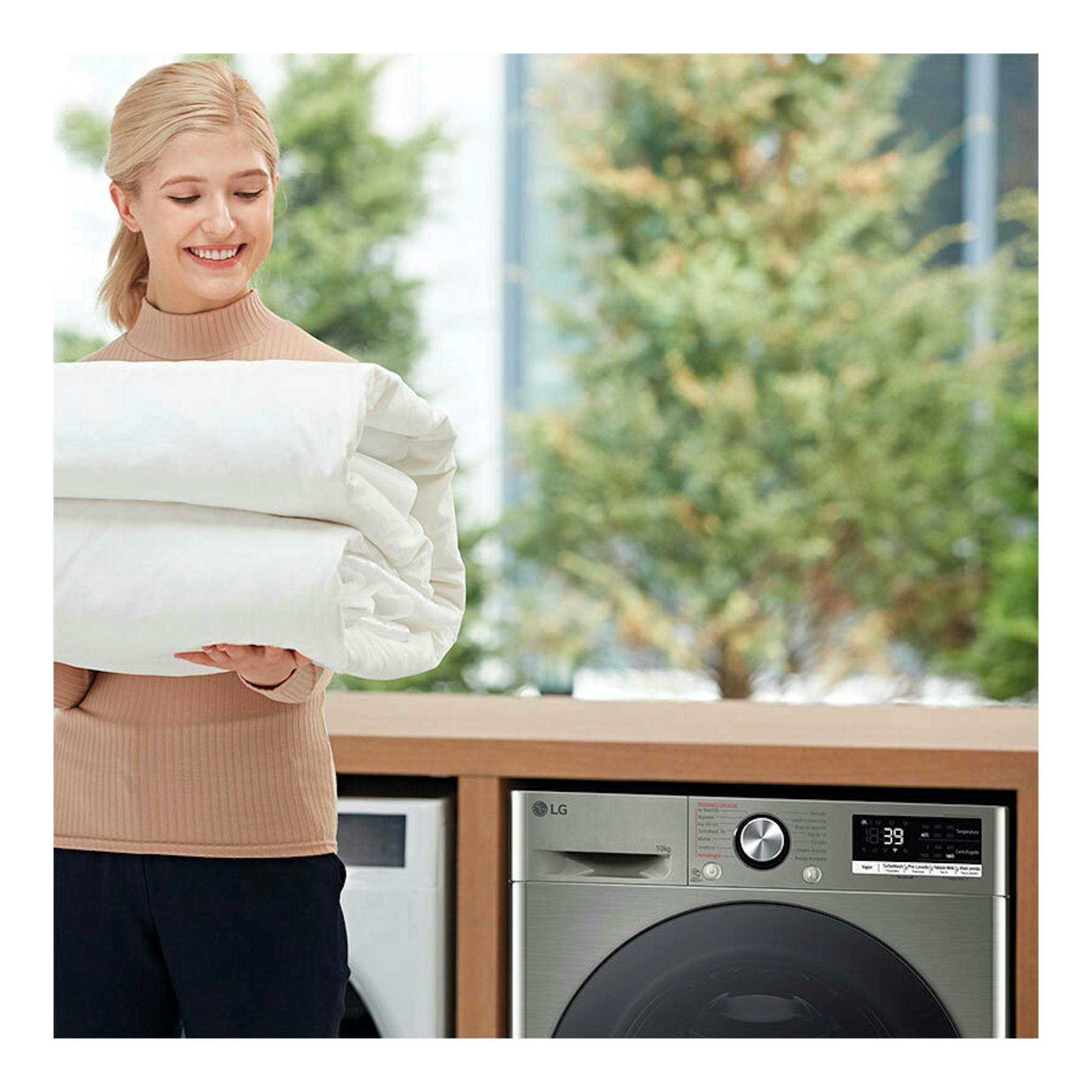 Lavadora LG F4WR7010AGS gris plata, vista frontal parcial, junto a una mujer con ropa de cama doblada.