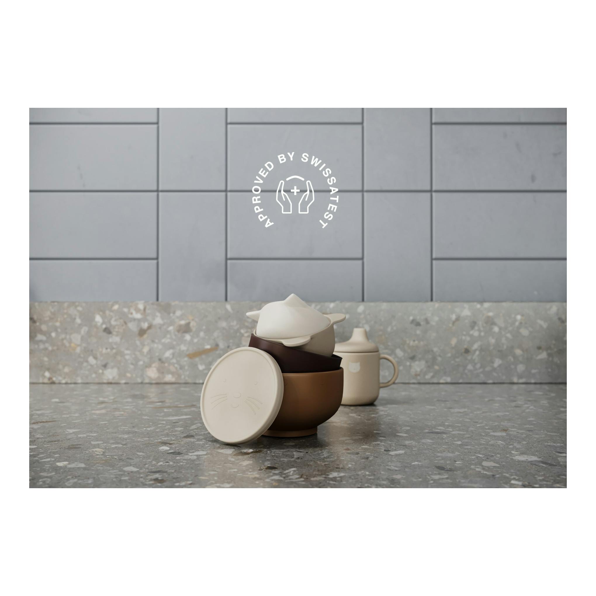 Vajilla apilada sobre encimera de cocina, con sello “Approved by Swissatest” al fondo.