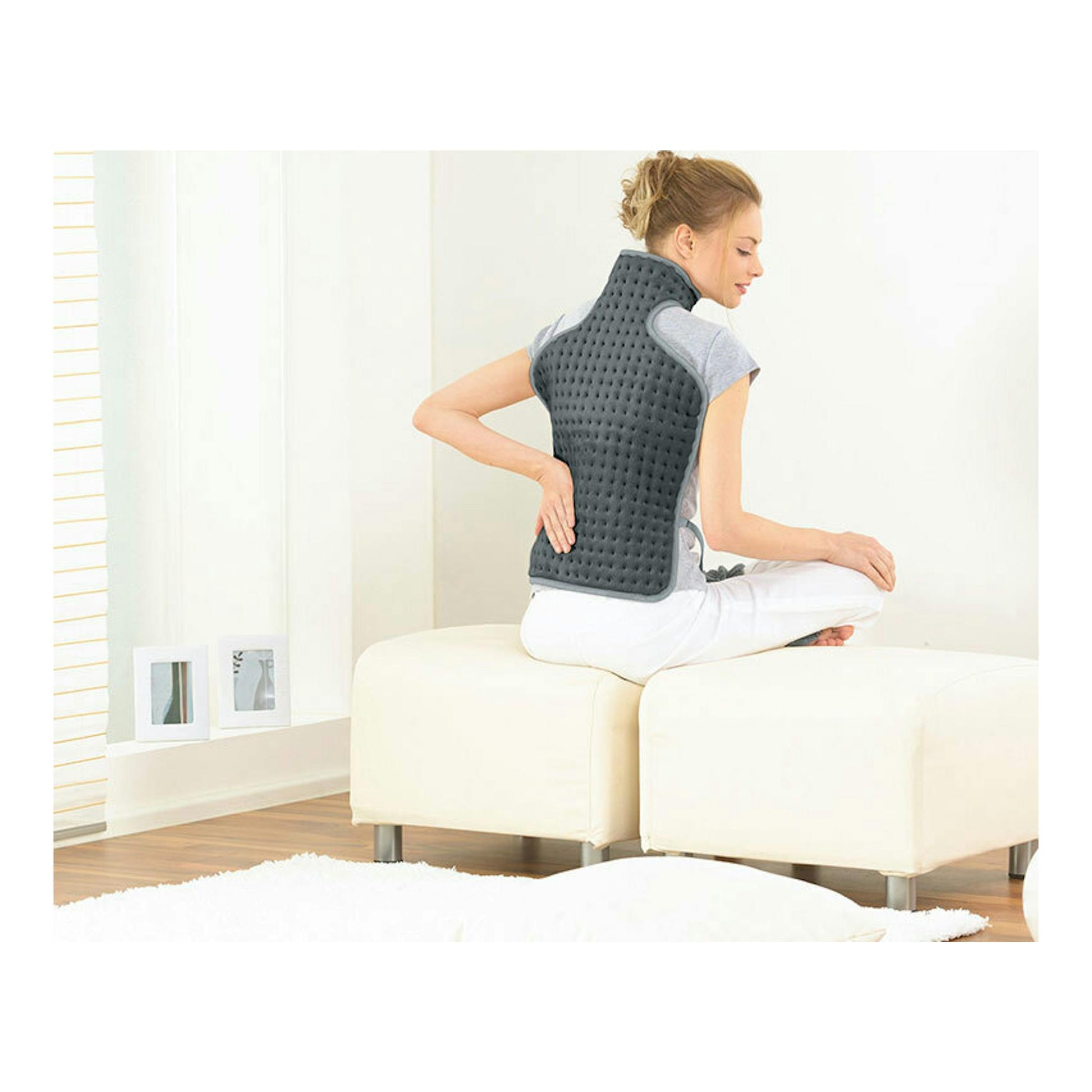 Manta eléctrica Beurer HK-53 gris colocada en la espalda de una mujer sentada de perfil sobre un asiento blanco.