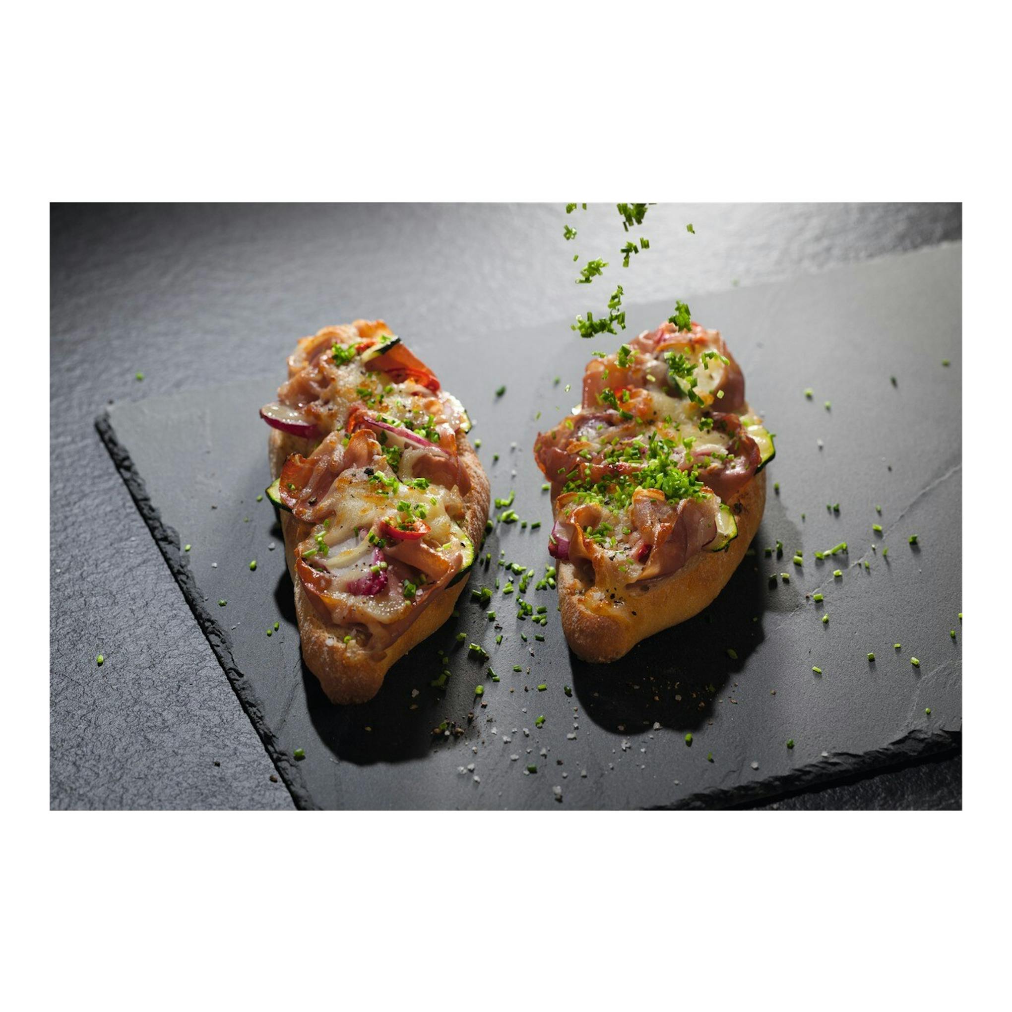 Dos tostas gratinadas con bacon, queso y verduras sobre pizarra negra, vista cenital.