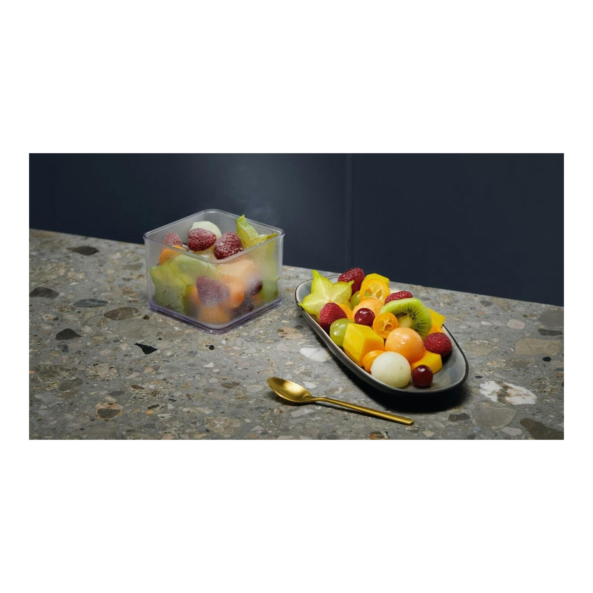 Fruta cortada en un recipiente y una bandeja sobre una encimera de piedra, con una cuchara dorada.