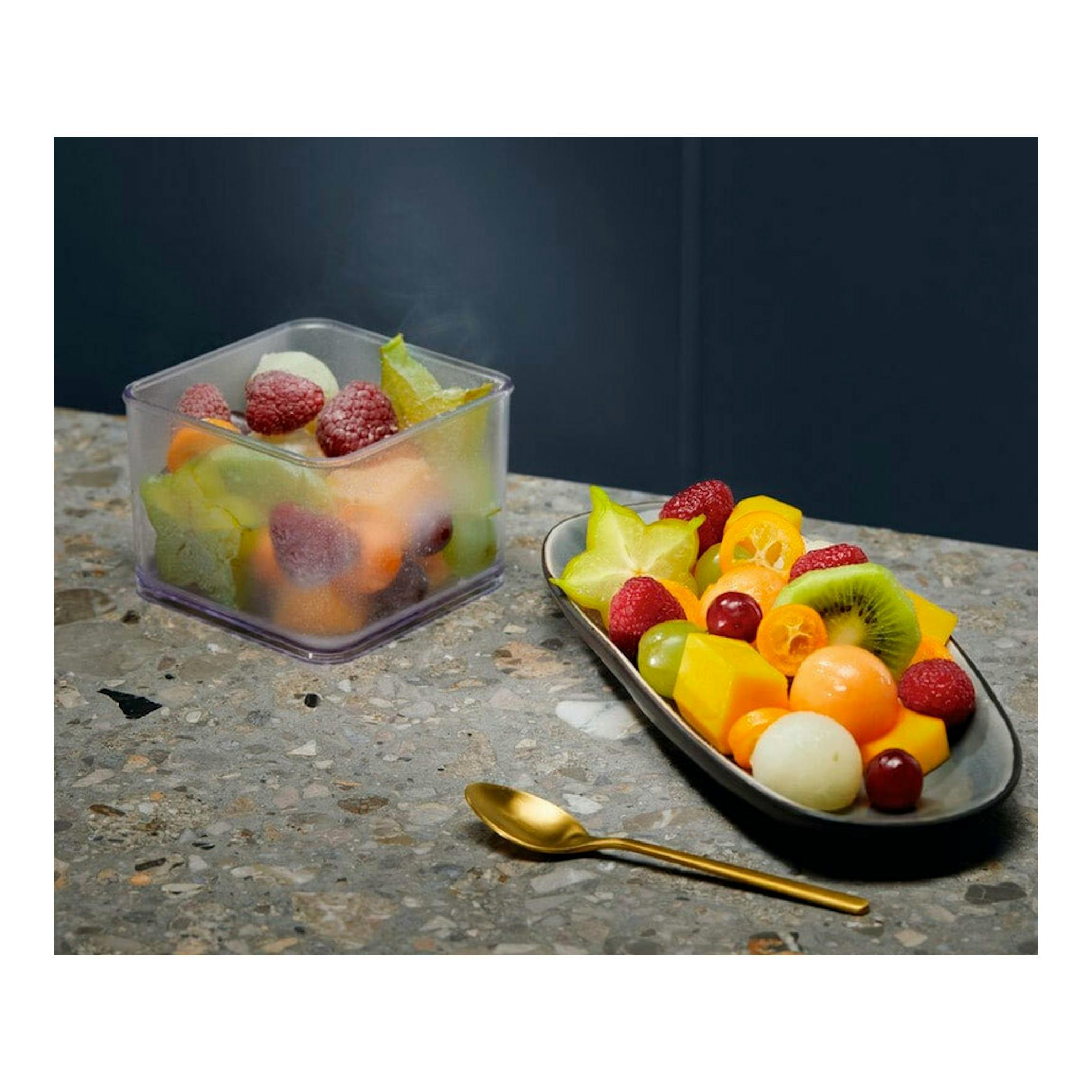 Recipiente transparente con fruta y plato ovalado con fruta cortada sobre superficie de piedra, y cuchara dorada.