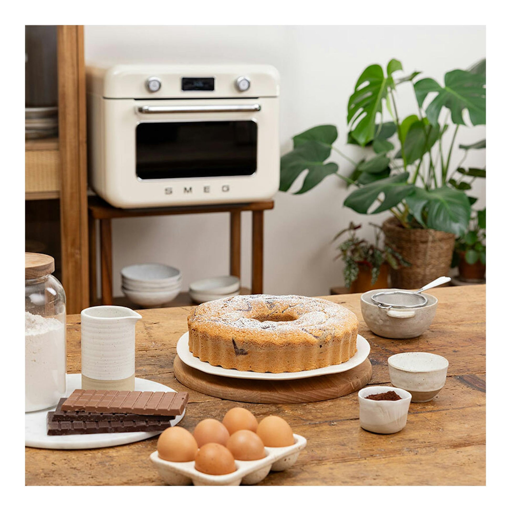 Minihorno Smeg COF01CREU crema, vista frontal en cocina junto a mesa con bizcocho e ingredientes.
