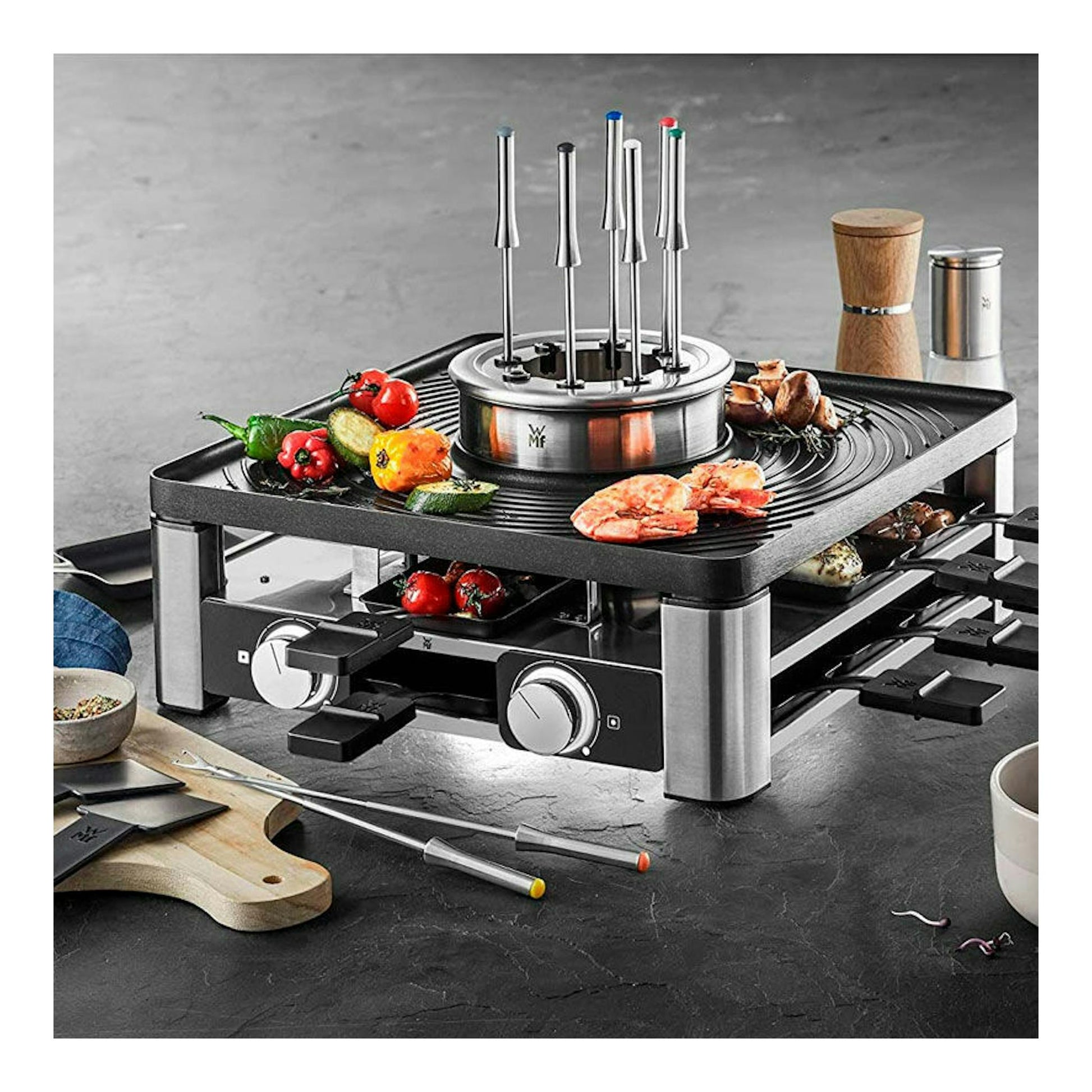 Parrilla grill WMF Gourmet de acero inoxidable y negro, vista 3/4, con fondue central y accesorios.