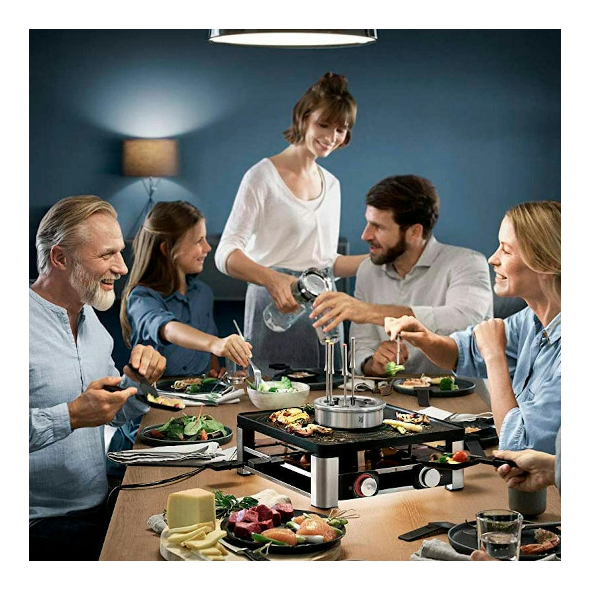 Parrilla de mesa WMF Gourmet negra y plateada en primer plano, sobre una mesa con comida y personas alrededor.