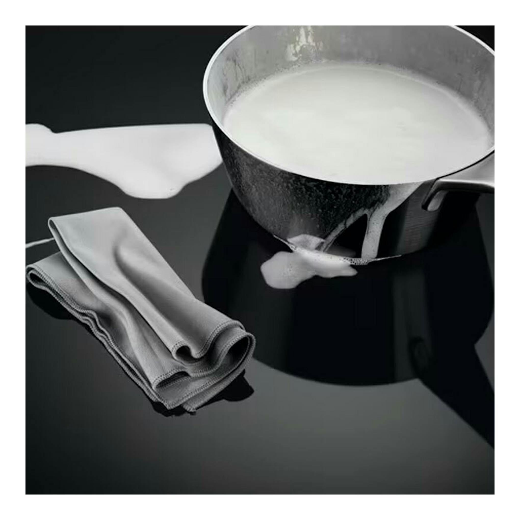 Superficie de placa de inducción negra con un cazo derramando leche y un paño gris doblado.