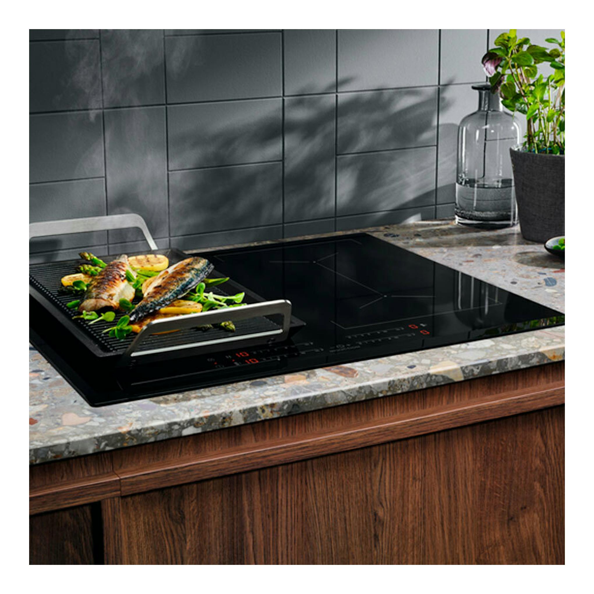 Placa de inducción Electrolux EIV644 negra, vista frontal, con bandeja grill y pescado en encimera.