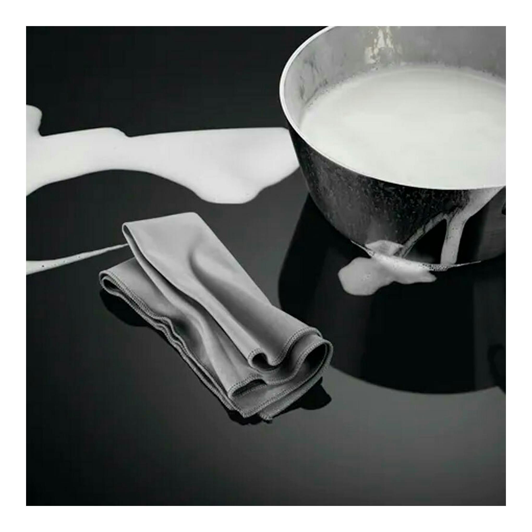 Placa de inducción AEG IKB63302FB negra, vista cenital parcial, con leche derramada, cazo y paño.